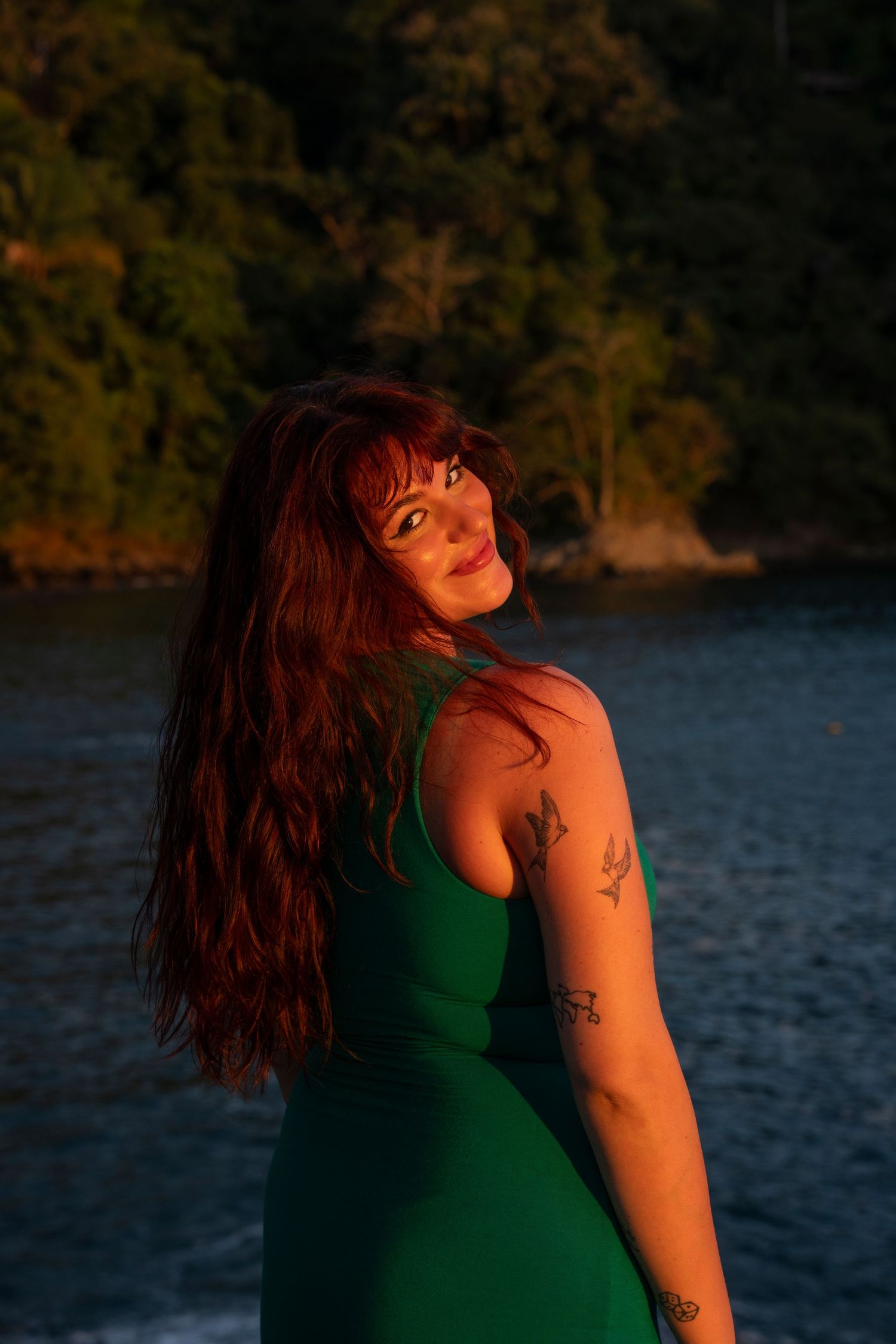 Woman with long red hair in green dress posing at sunset near water.