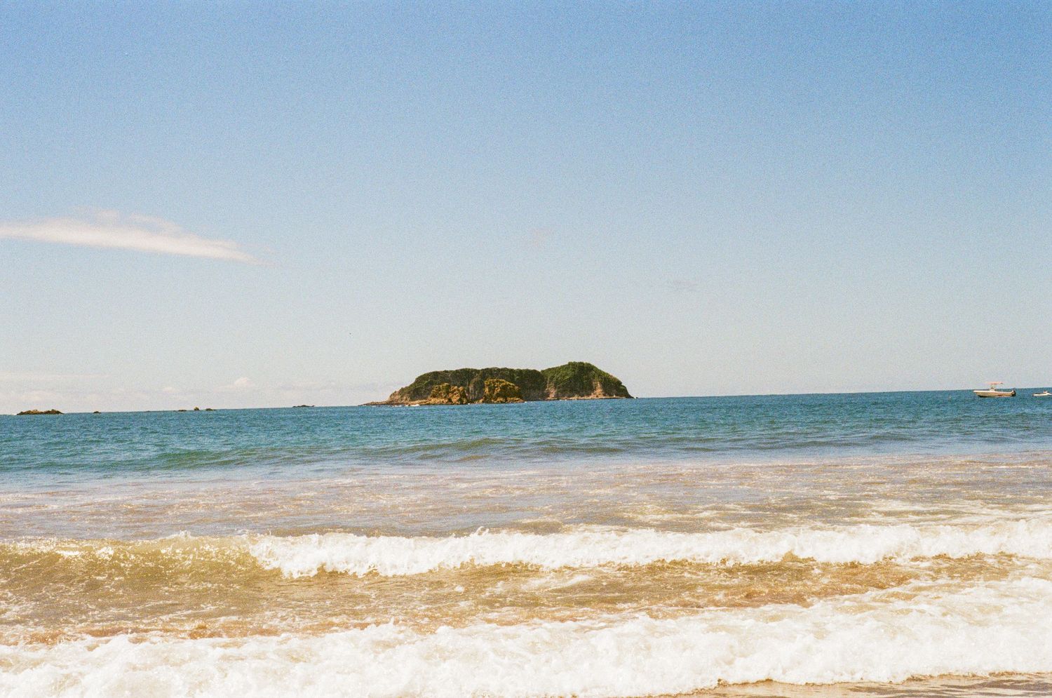 Small rocky island surrounded by calm blue ocean waters viewed from a sandy beach with gentle waves.