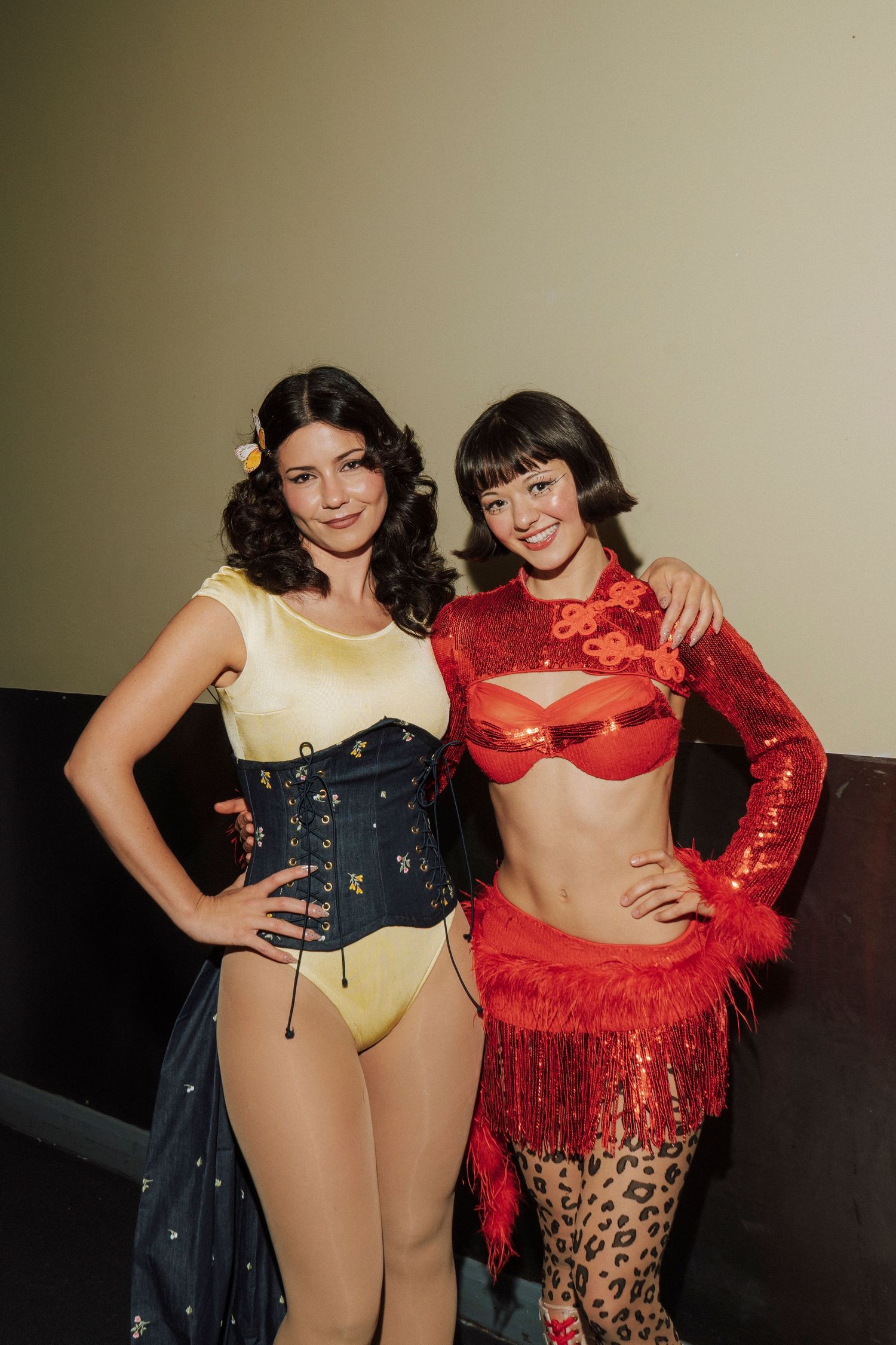 Two women in colorful performance costumes pose together, one in yellow and black corset, other in red sequined outfit.