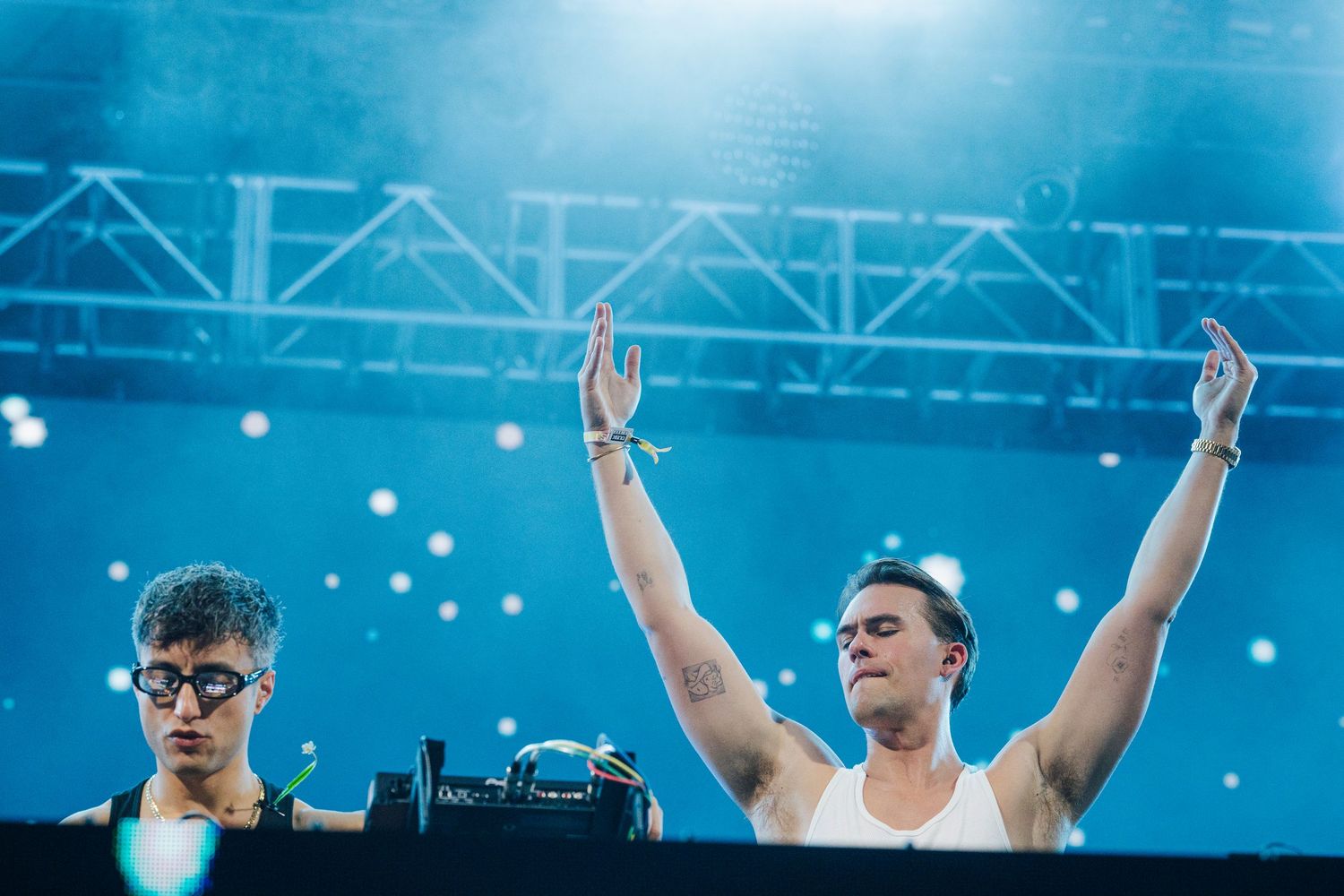 Two male performers at DJ booth on stage with blue atmospheric lighting and raised hands.
