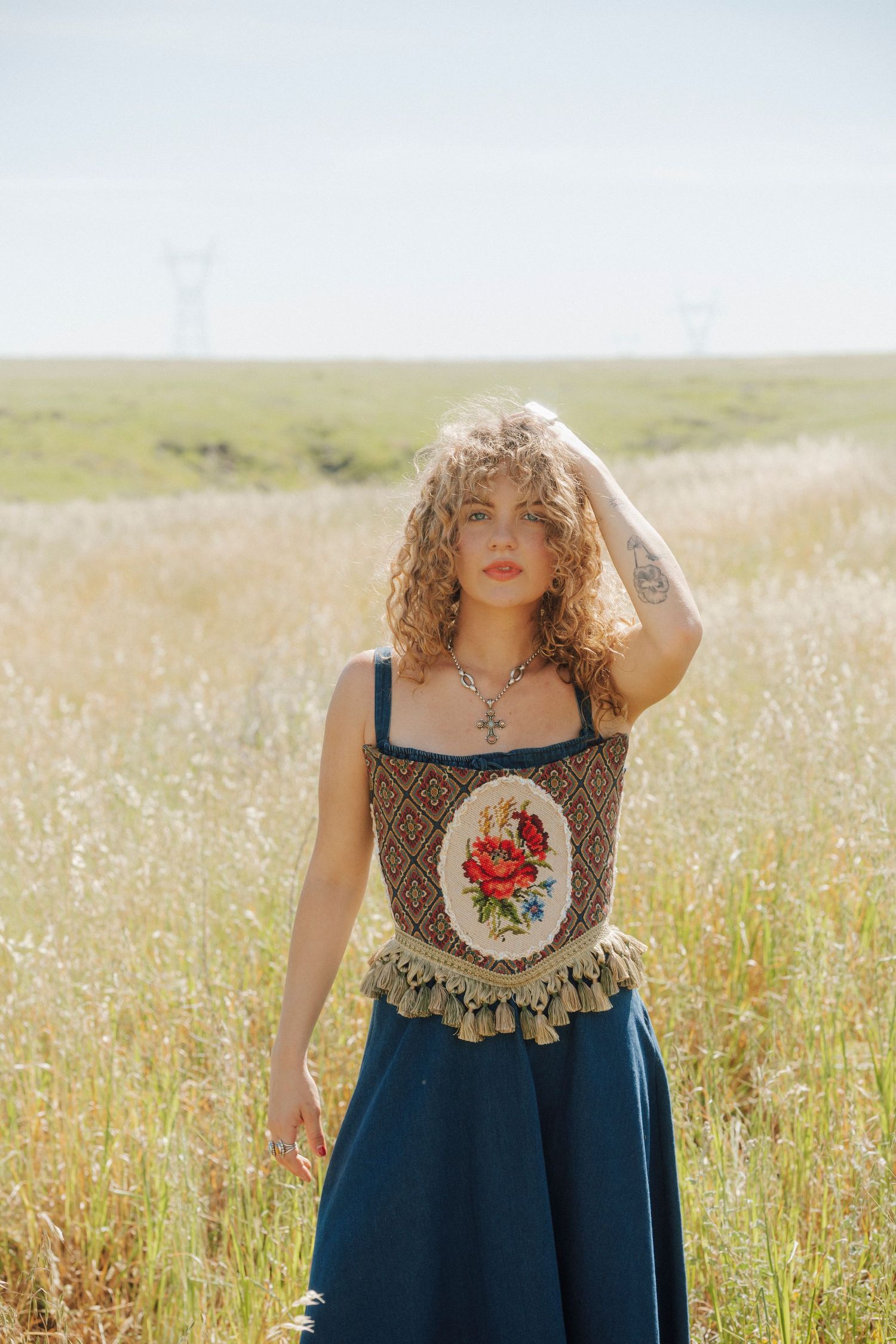 Curly-haired woman in embroidered floral corset and blue skirt standing in a sunny golden field.