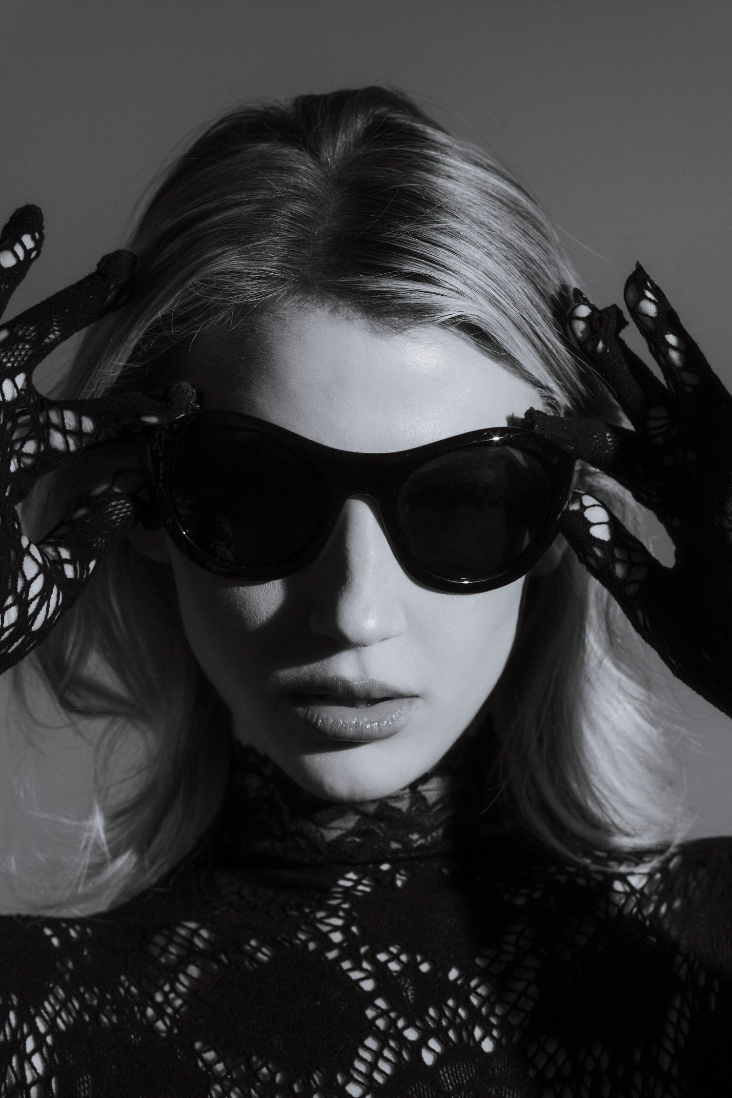 Black and white portrait of blonde woman wearing oversized sunglasses and lace gloves in editorial fashion style.