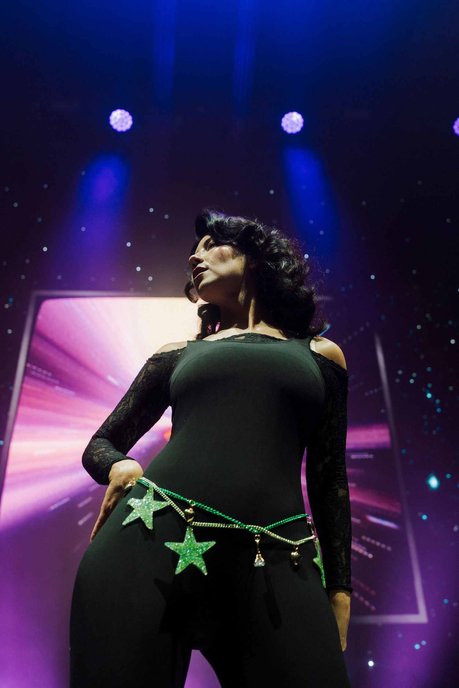 Performer in black bodysuit with green star belt poses on stage under blue and purple spotlights.