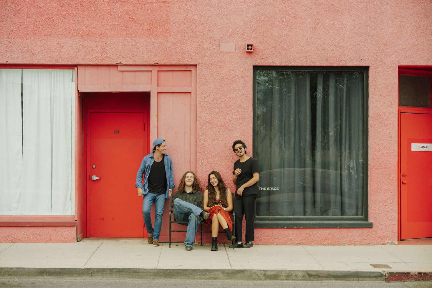Four people posing casually against a pink building with red doors on a city sidewalk.