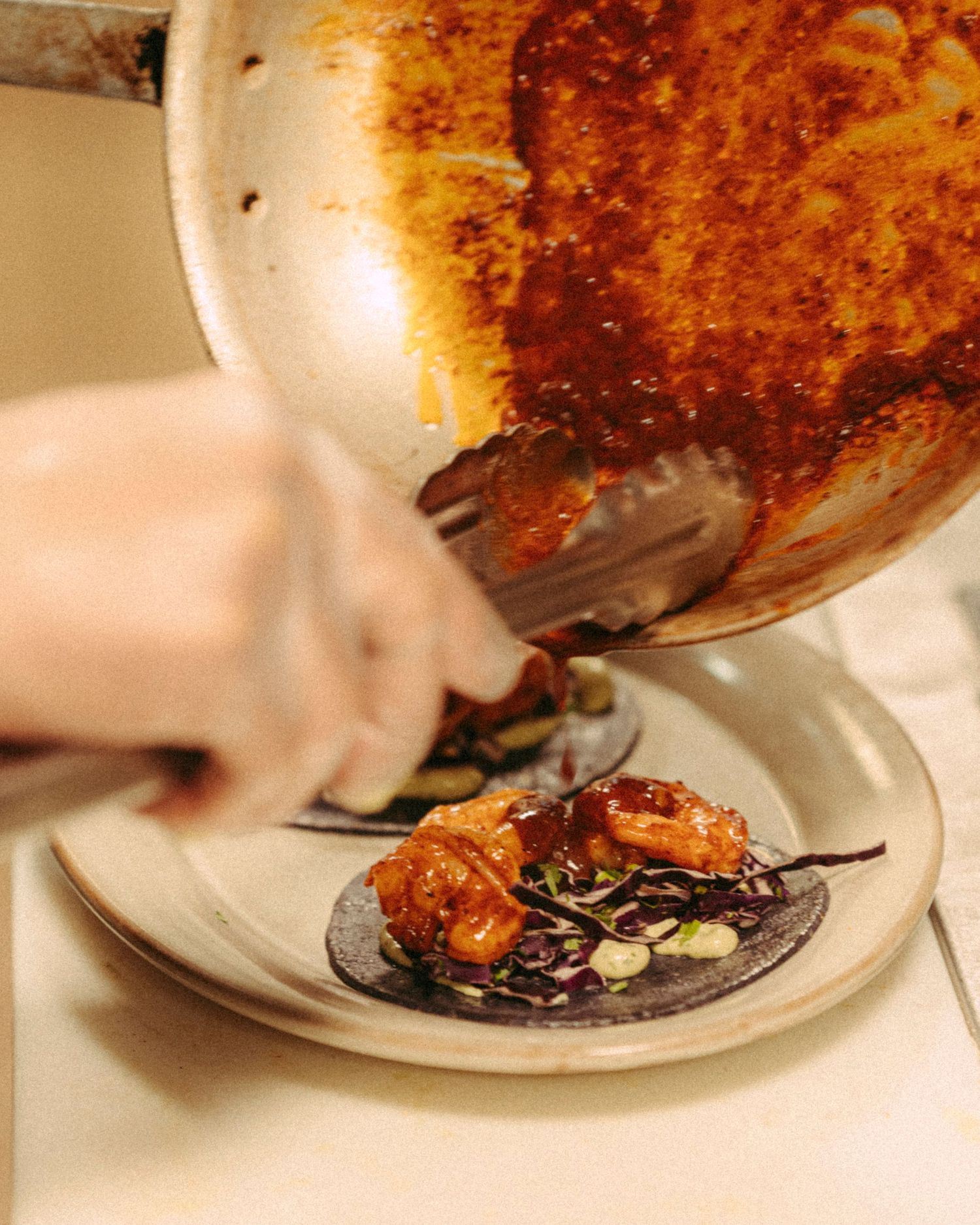 Chef plating shrimp tacos with sauce from a pan onto a dark tortilla.