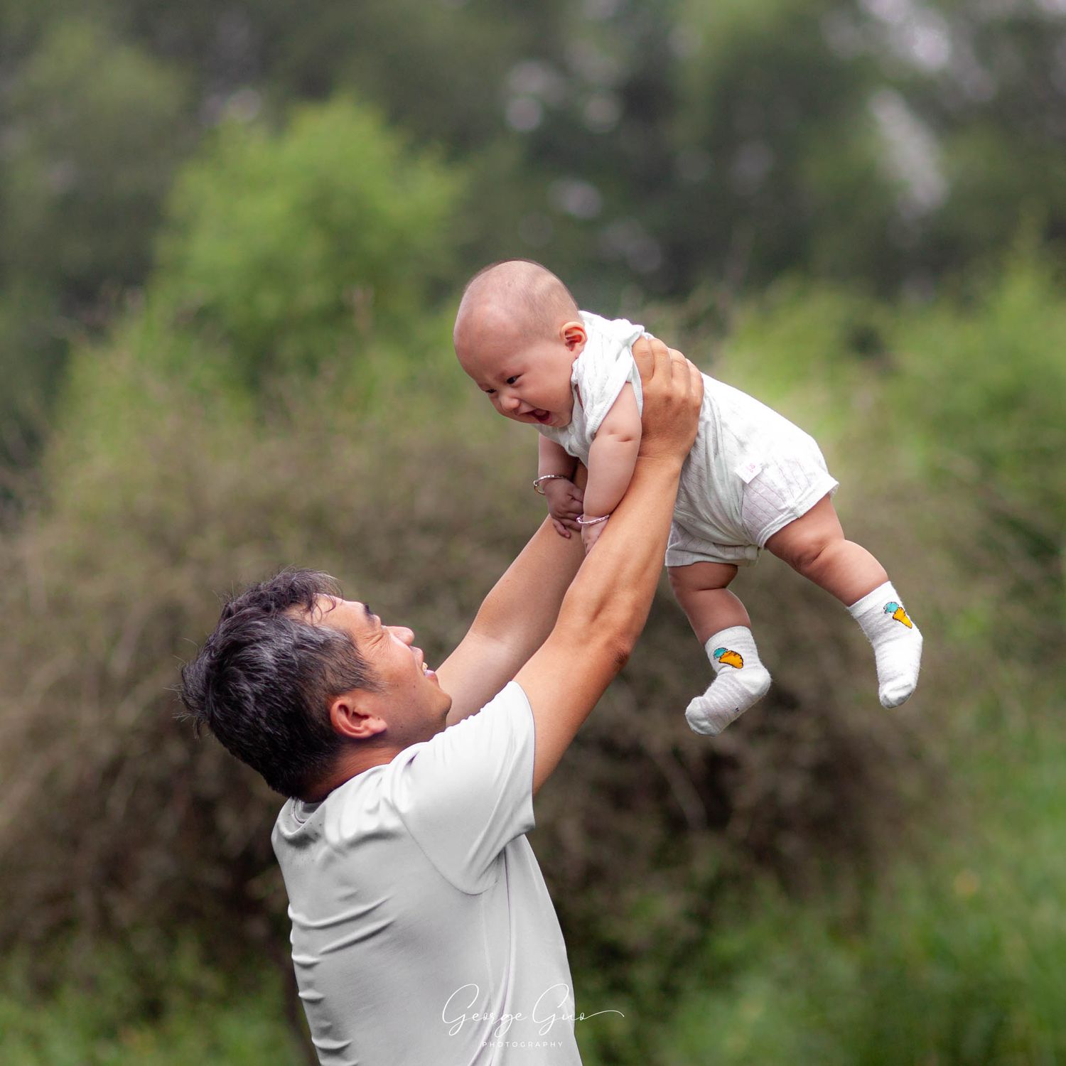 A man lifting and holding his infant child in the air, playing happily and joyfully in a green park, photographed by George Guo Photographer