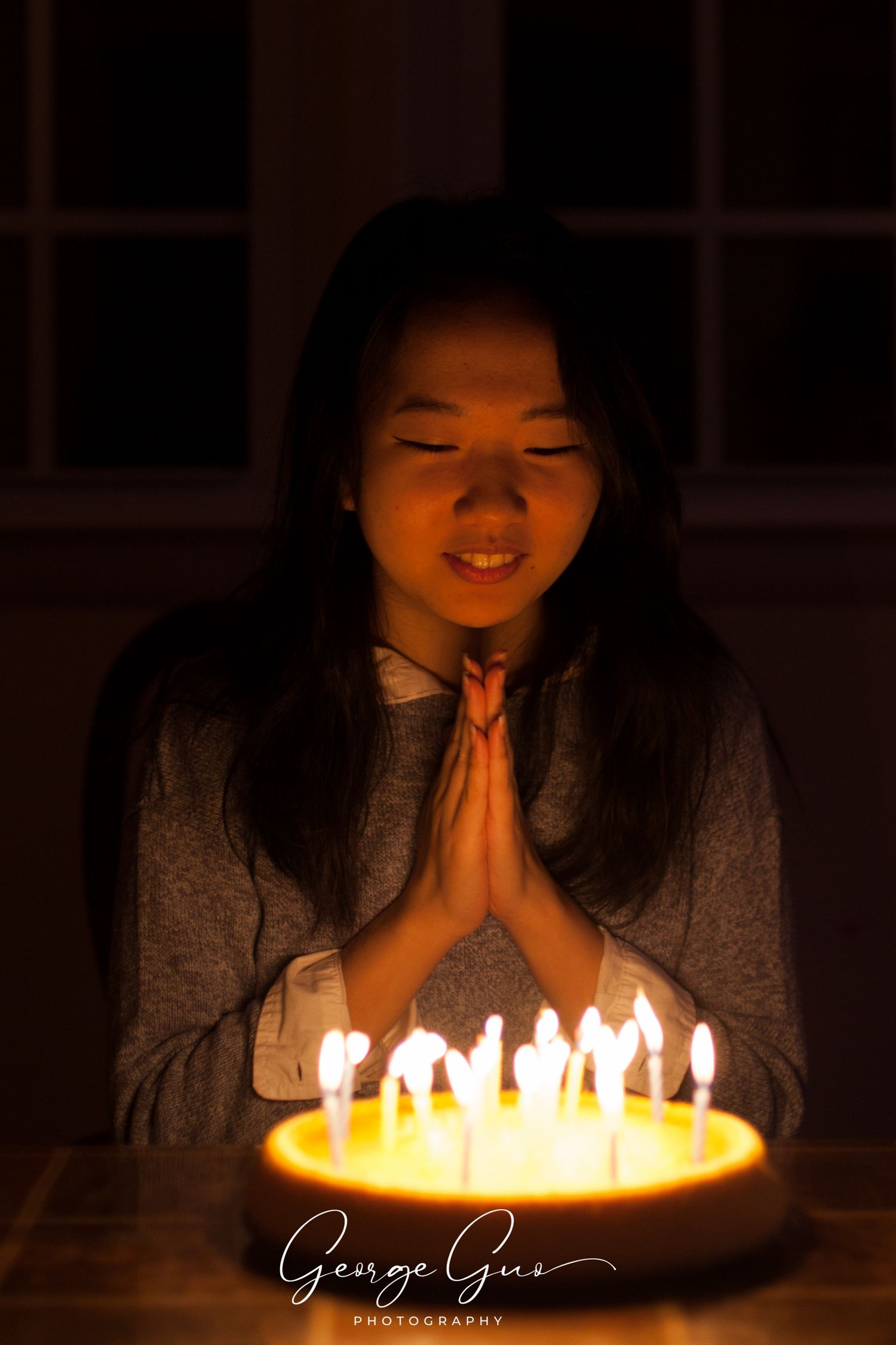 A young girl praying and making a wish before blowing out candles on a birthday cake, captured in low-light natural ambiance by George Guo