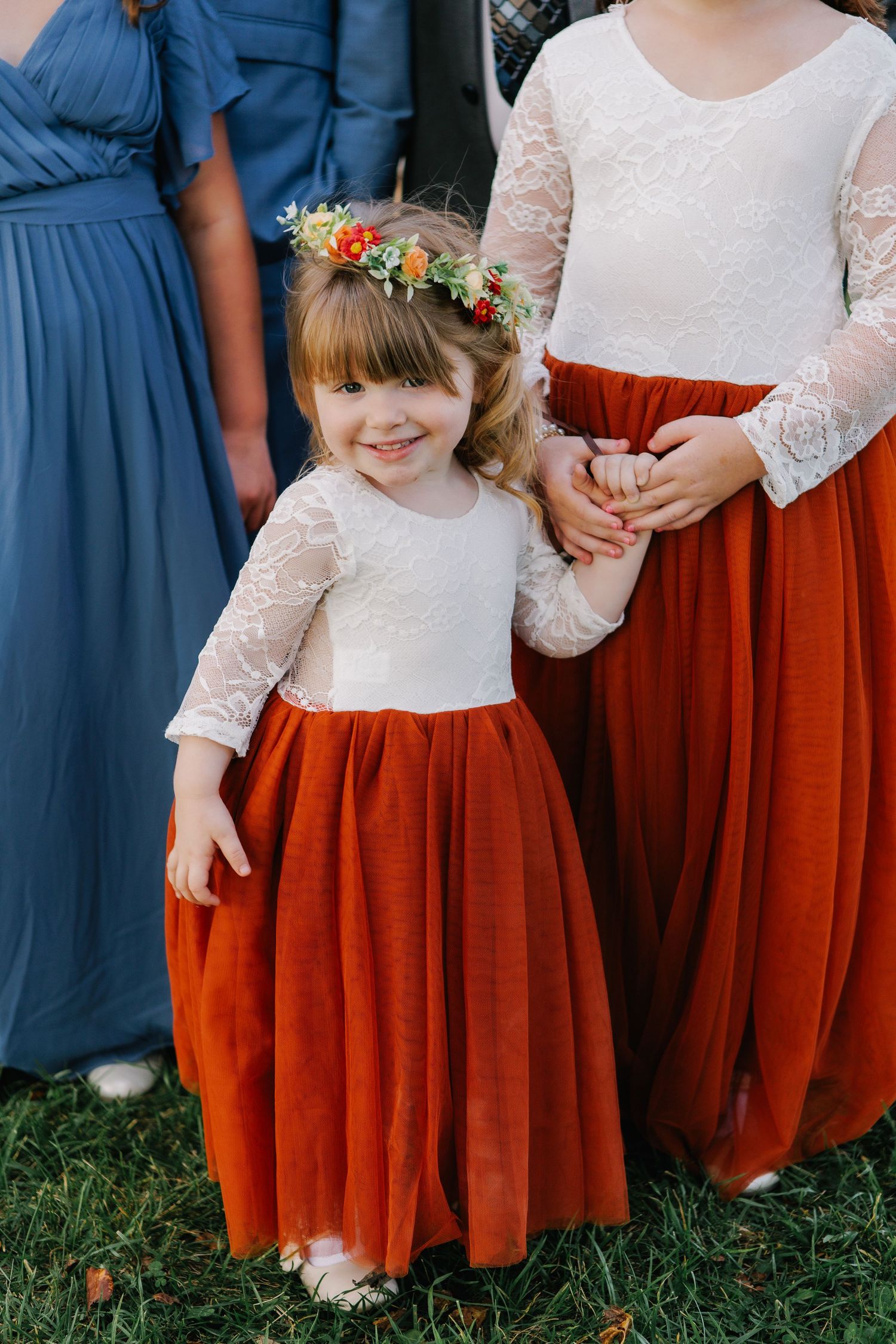 Bridesmaids in navy blue dresses and flower girls in white lace tops with rust-colored skirts stand together outdoors.