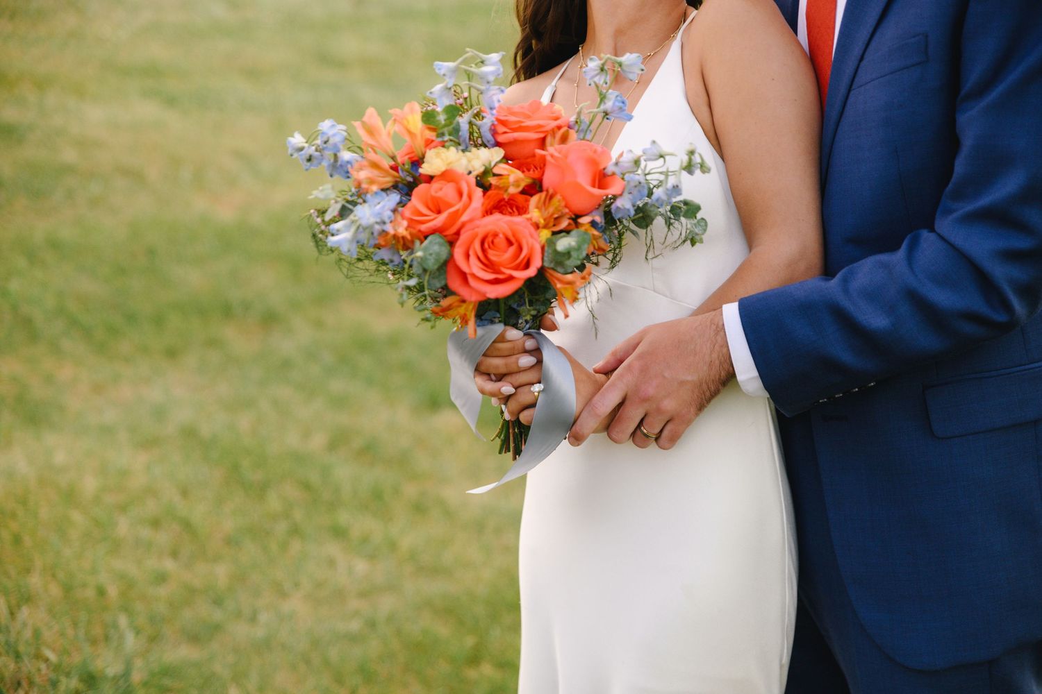 Bride and groom holding a vibrant orange rose and blue wildflower wedding bouquet outdoors on a green lawn.