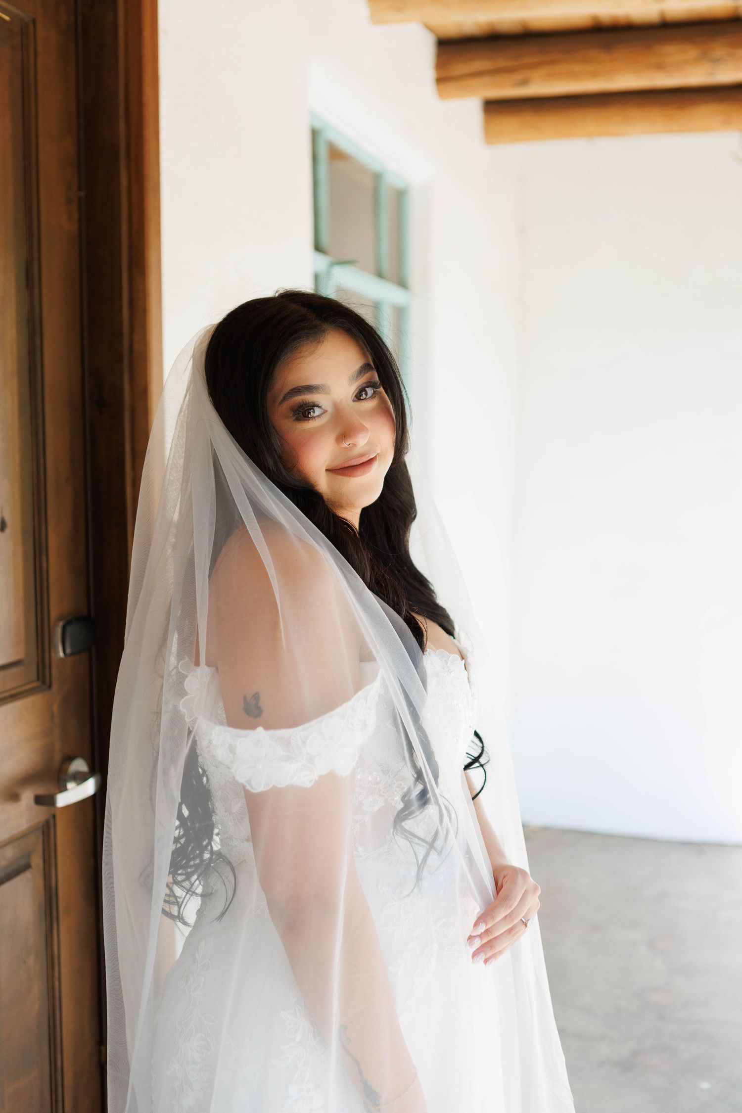 Bride in white lace gown and sheer veil poses elegantly by a wooden door in a sunlit venue.