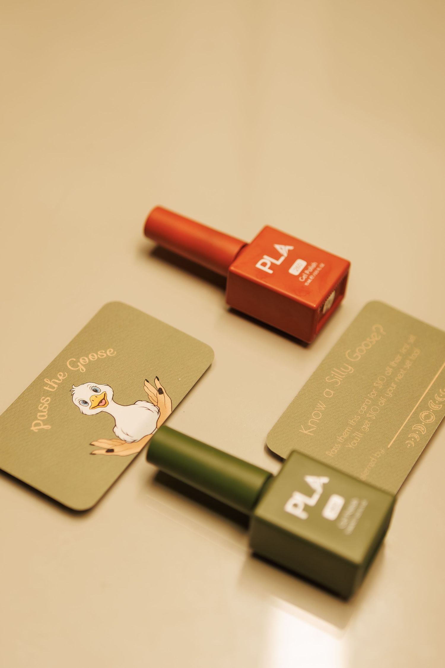Two PLA gel nail polish bottles in red and green beside cards featuring a goose illustration on a beige surface.