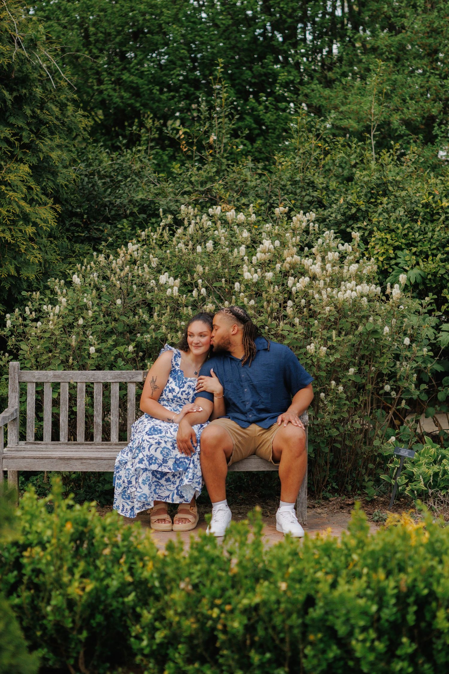 Couple sitting on wooden garden bench, man kissing woman's cheek, surrounded by lush green shrubs and white flowering bushes.
