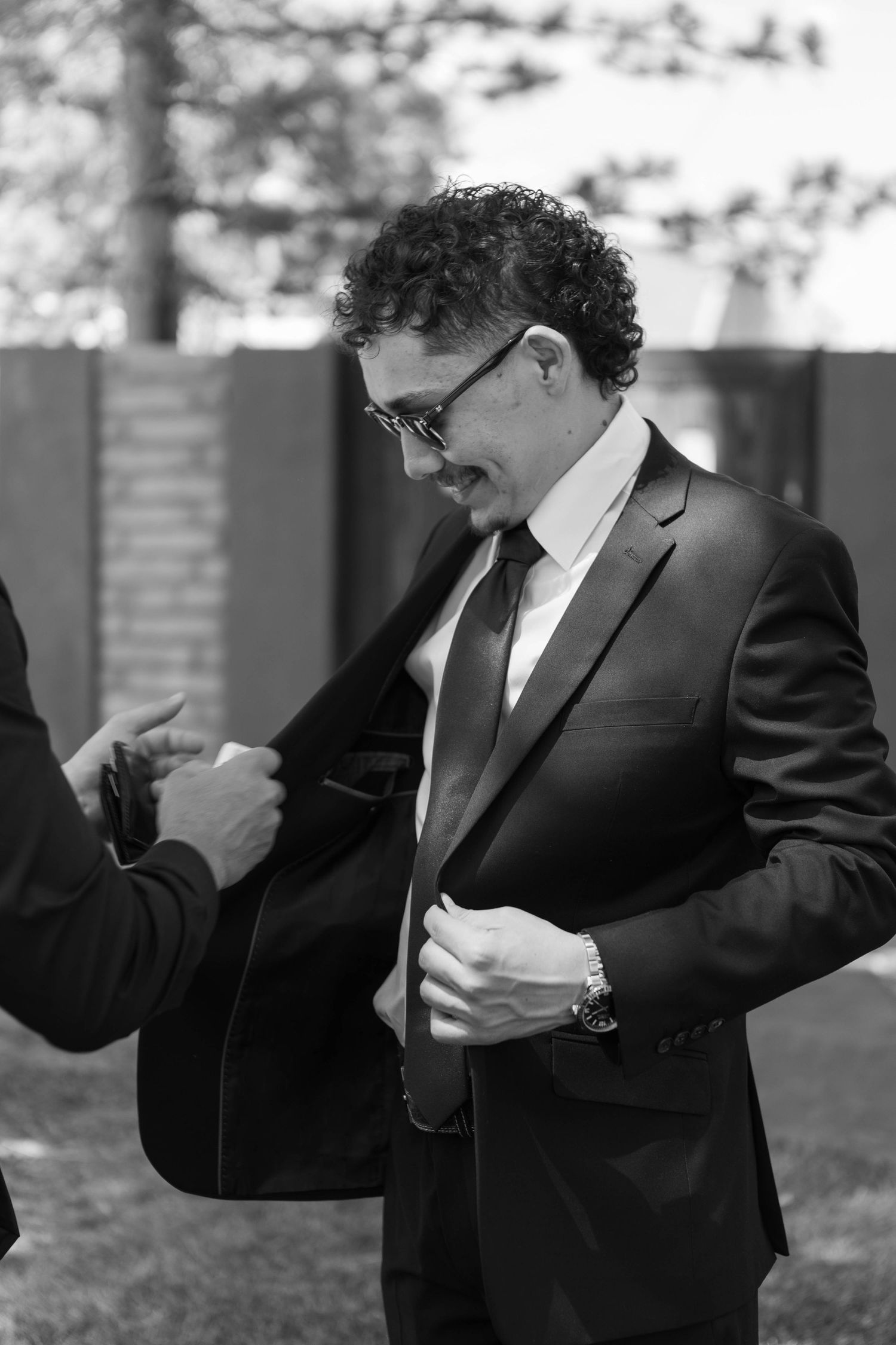 Black and white photo of a smiling man in a suit and glasses adjusting his jacket outdoors.