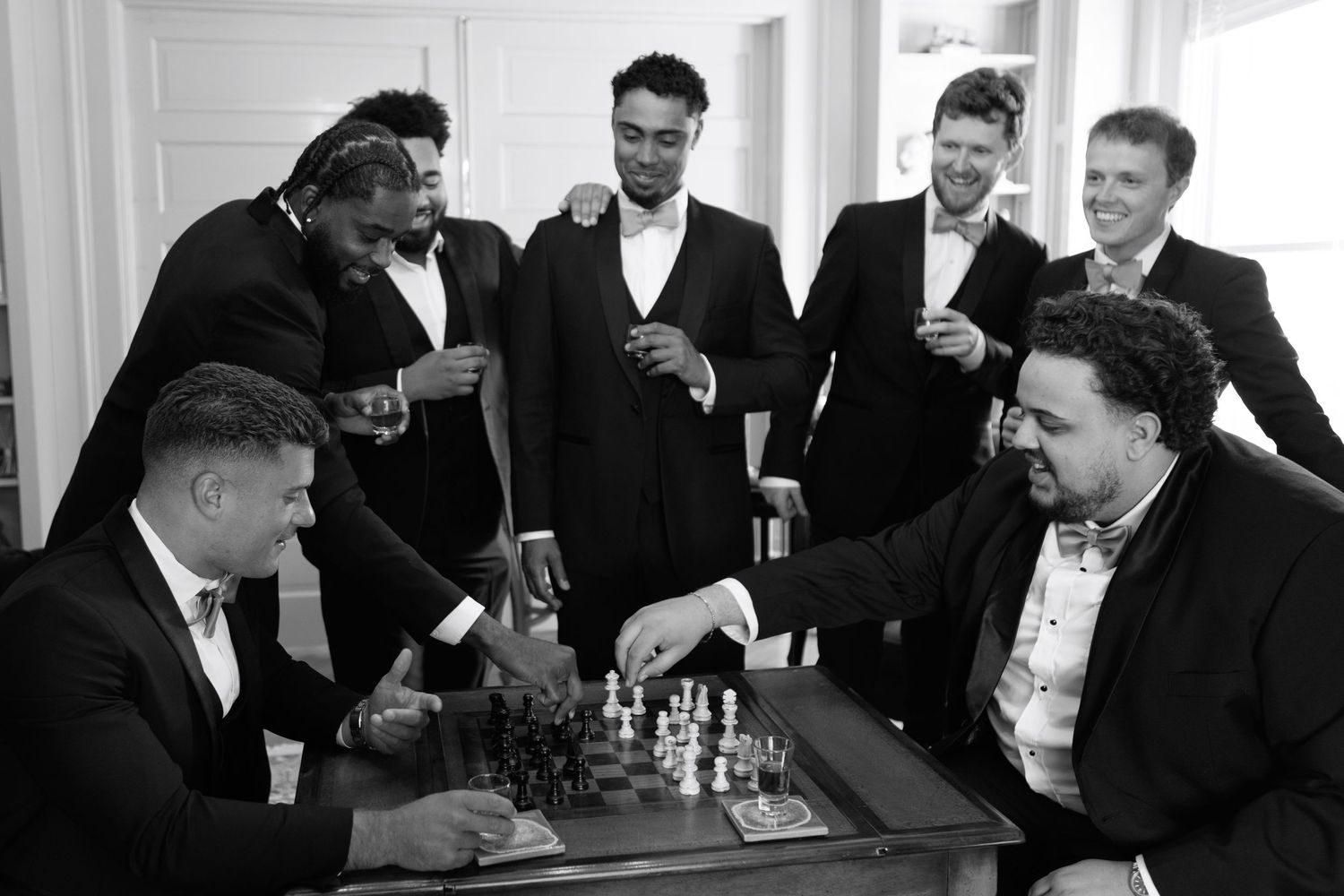 A group of men in suits laugh and smile while playing chess at a social gathering.