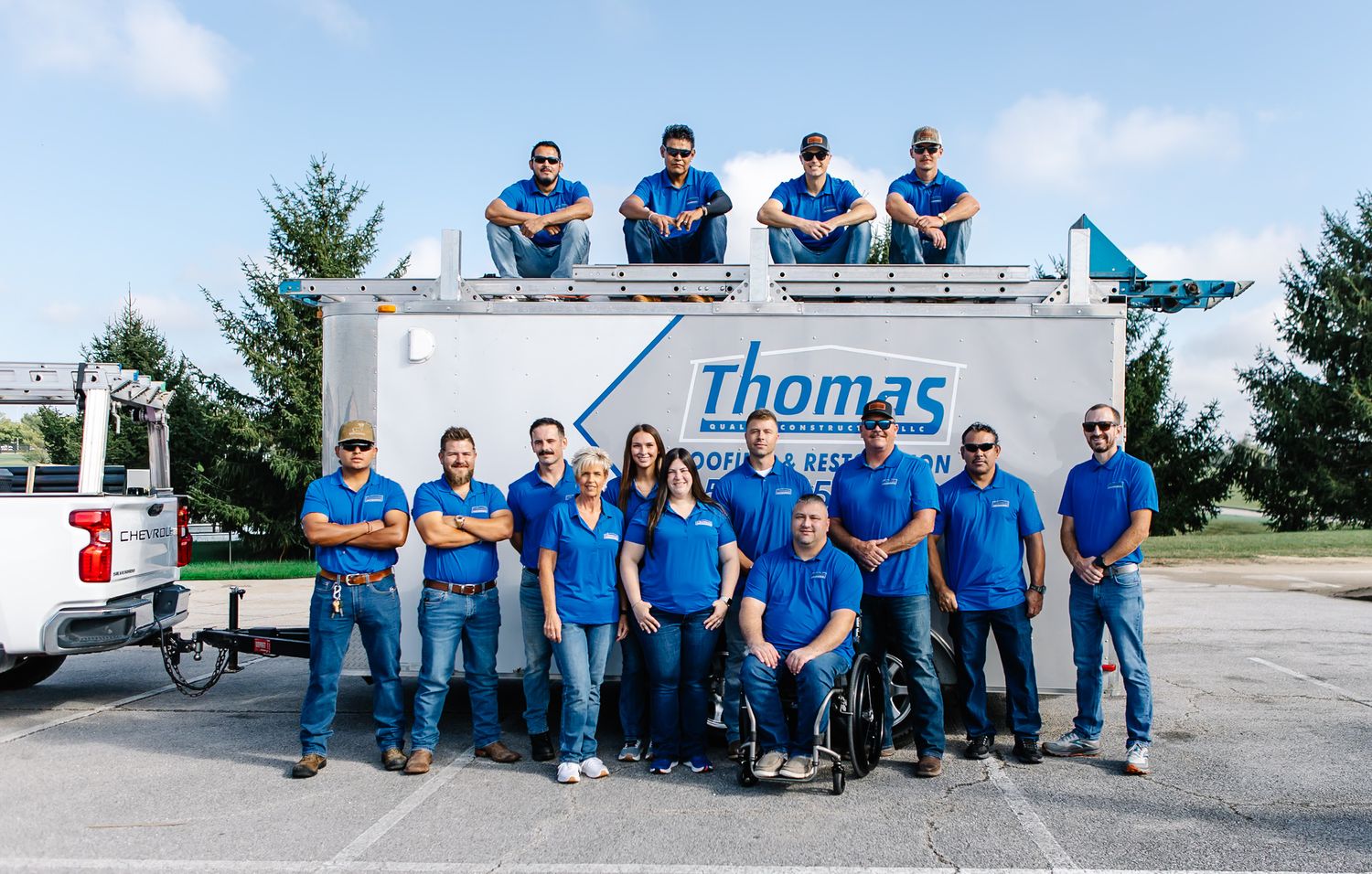 Group of workers in blue uniforms standing in front of Thomas Roofing company vehicles.