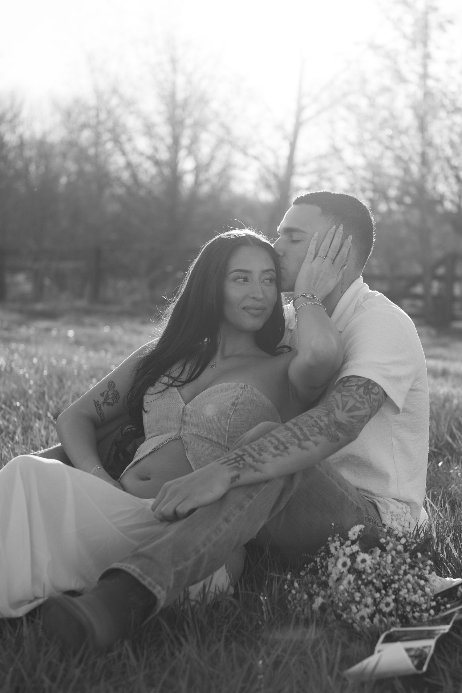 Black and white photo of a couple sitting on grass, man kissing woman's cheek, both with tattoos, trees in background.
