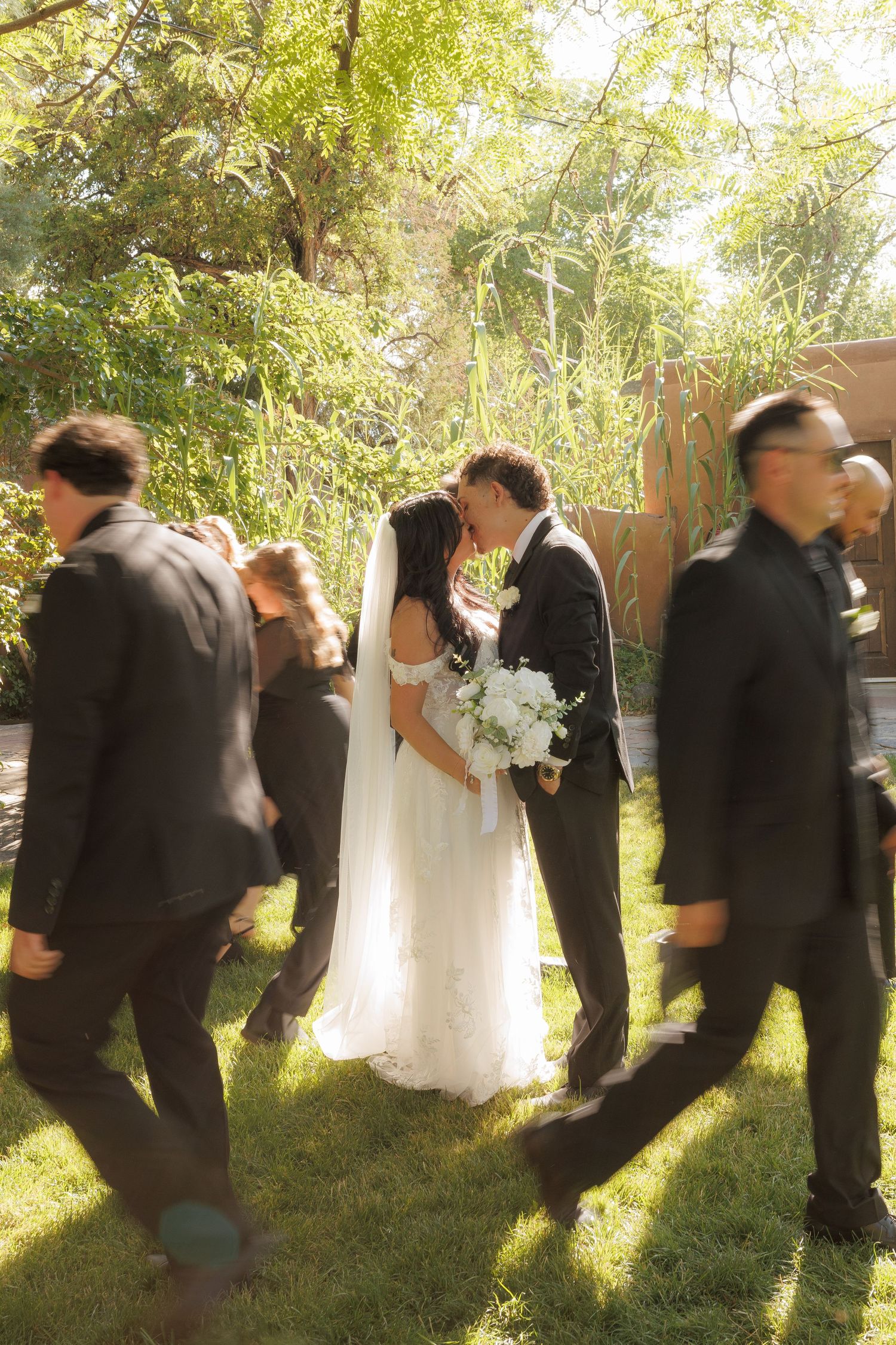 A bride and groom share a kiss outdoors on green grass, surrounded by guests in dark suits, with lush trees in the background.