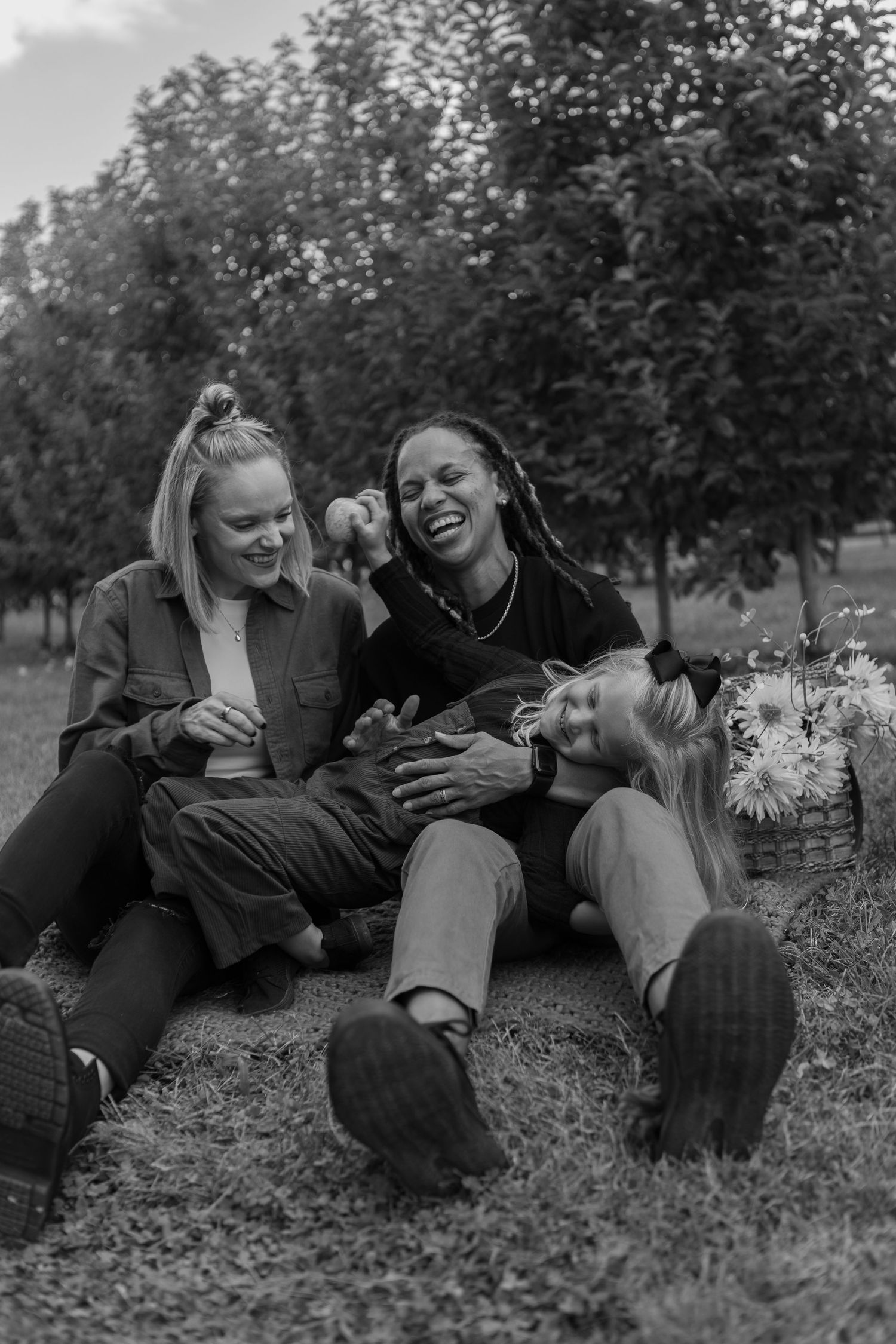 Three women laughing and sitting together on grass in a park, enjoying a picnic in black and white photo.
