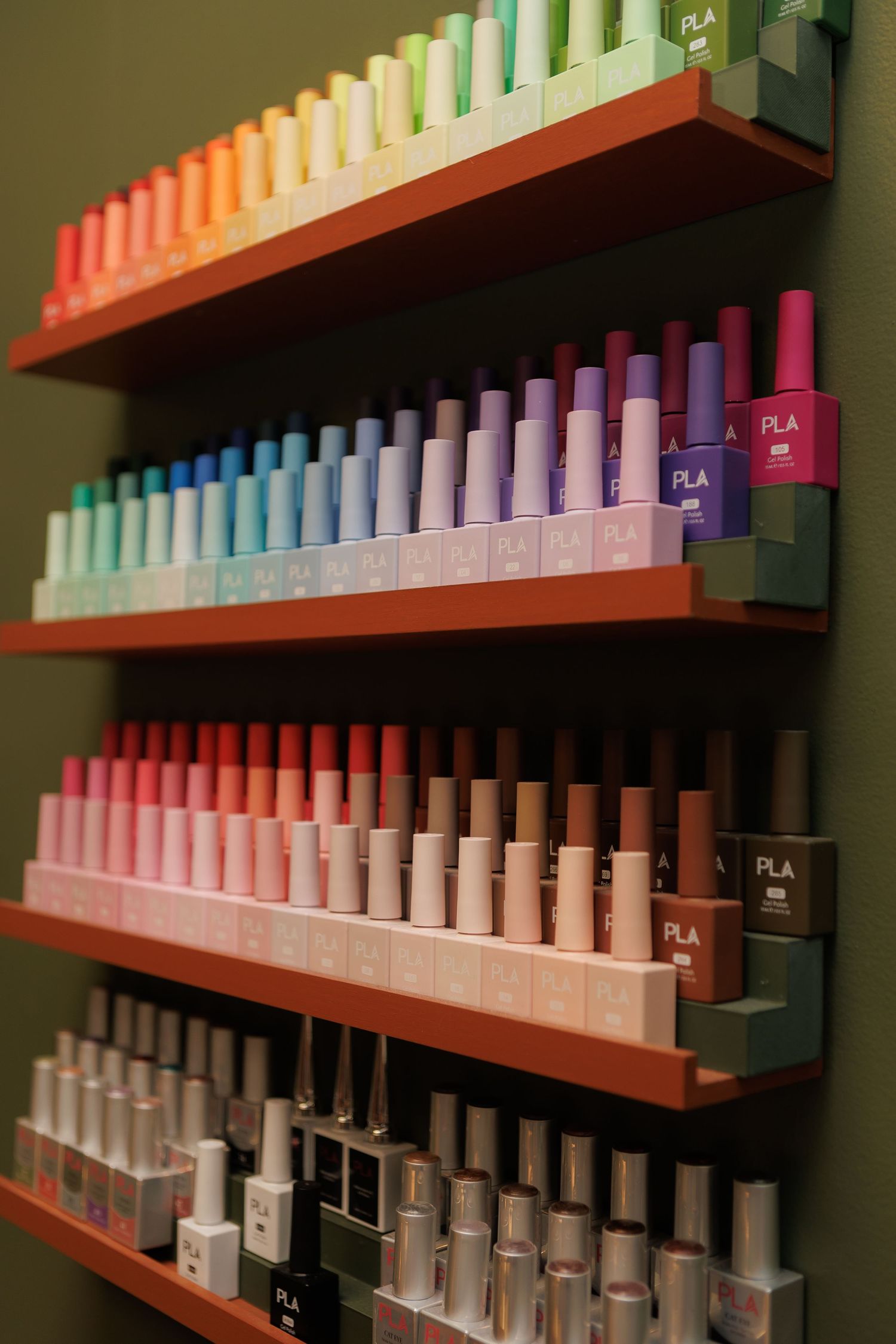 Colorful PLA nail polish bottles arranged by color on orange wall-mounted shelves in a beauty salon.