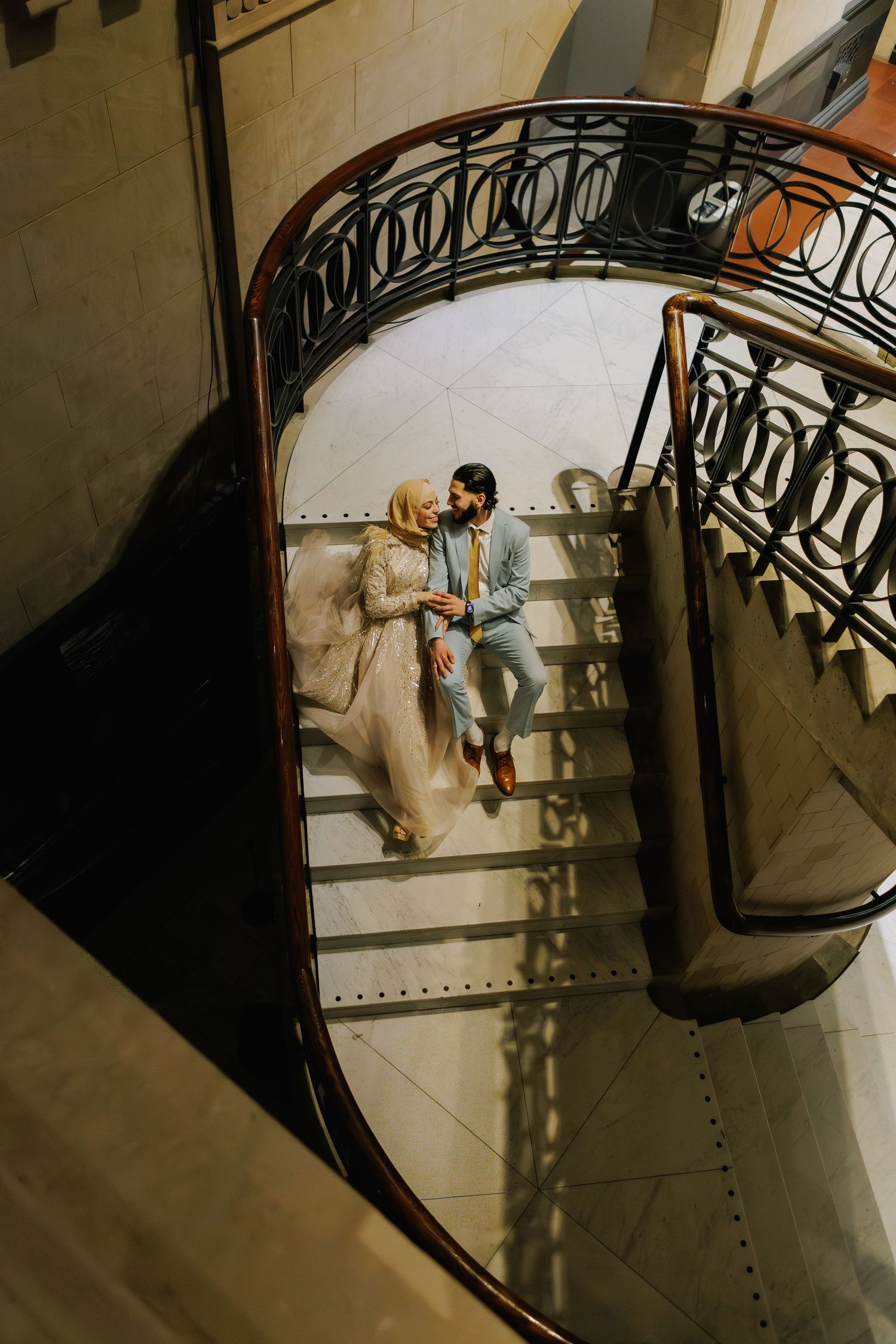 Aerial view of a couple in elegant attire sitting on a grand spiral staircase with ornate iron railings.