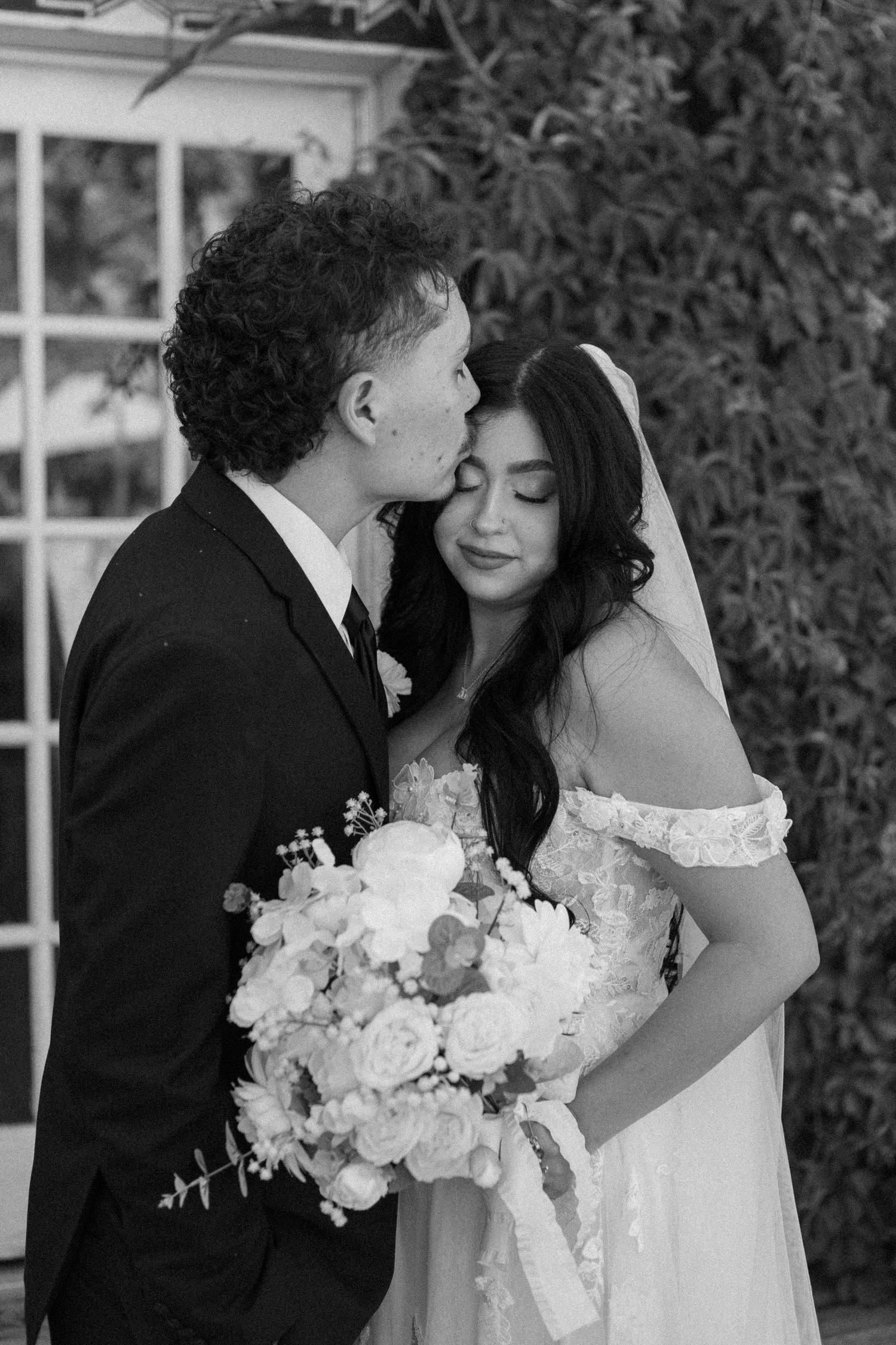 Black and white photo of a groom kissing his bride on the cheek as she holds a lush white floral bouquet outdoors.