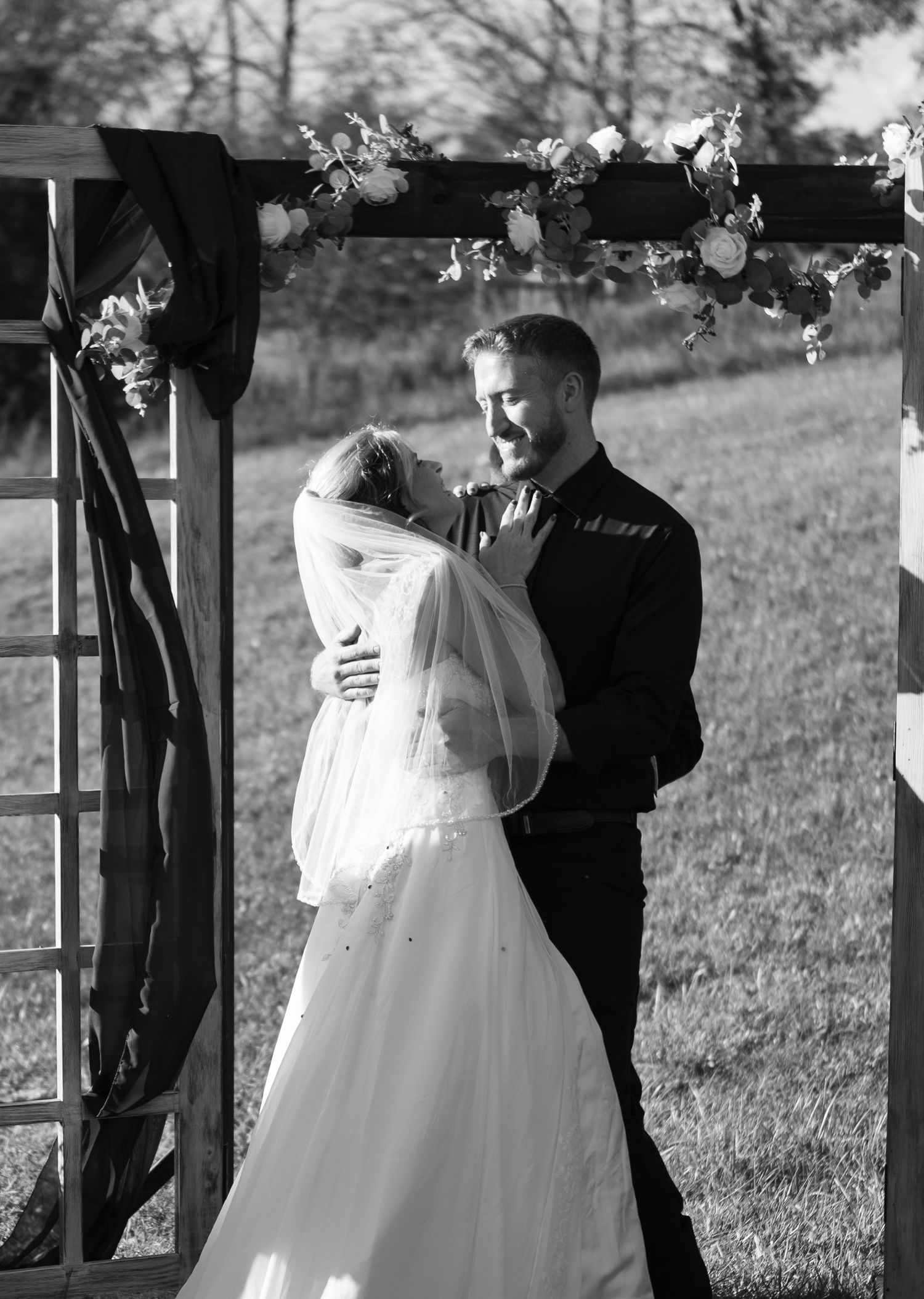 Black and white photo of a wedding ceremony taking place under a wooden arch outdoors.