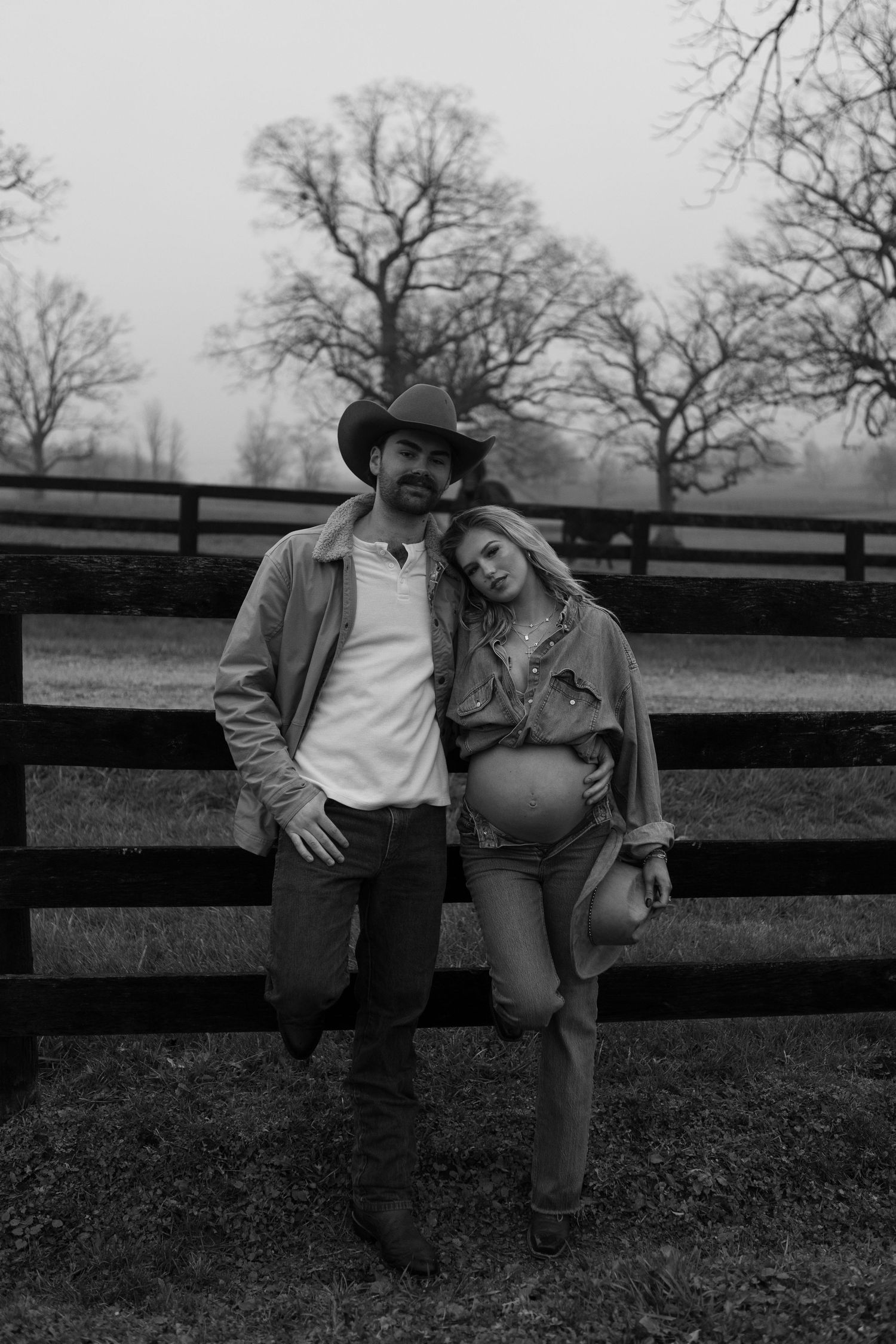 Couple sits together on wooden fence with bare trees in background.