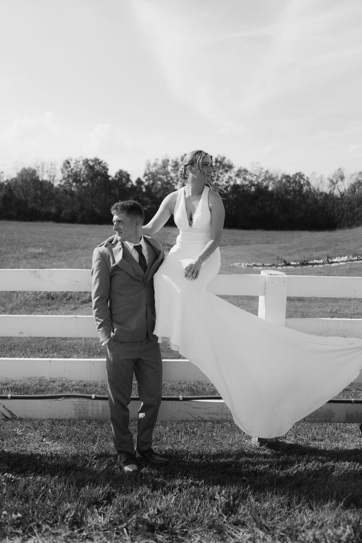 Black and white wedding photo with flowing dress caught in wind by white fence.
