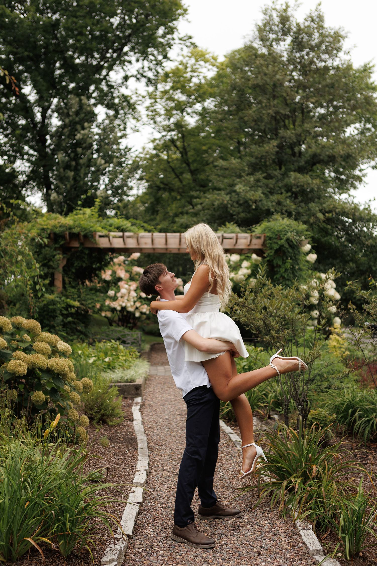 A man lifts a woman in a white dress on a garden path surrounded by lush greenery and blooming flowers.