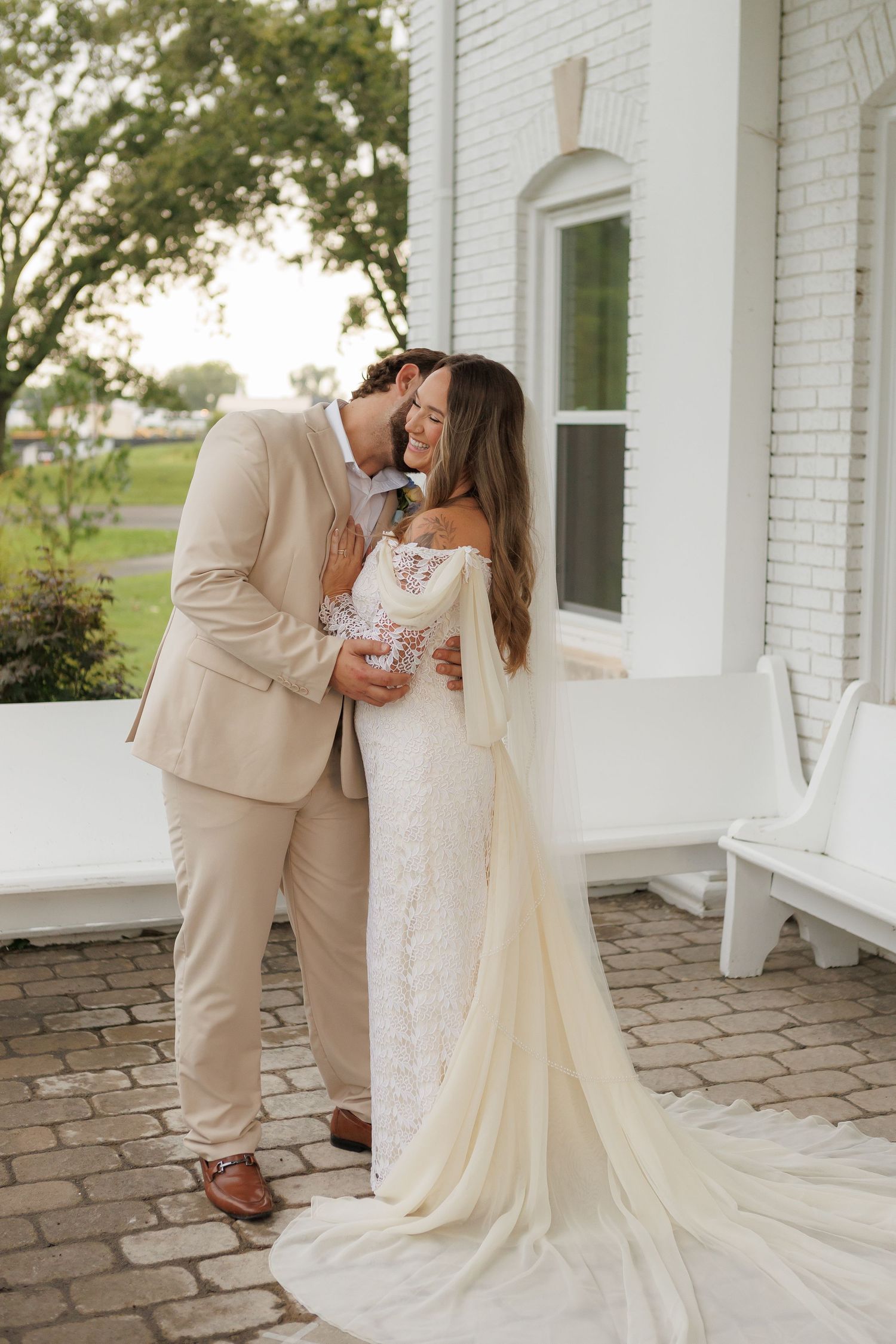 A bride and groom share a romantic kiss outside a white brick chapel, she wearing a lace gown with cape sleeves.