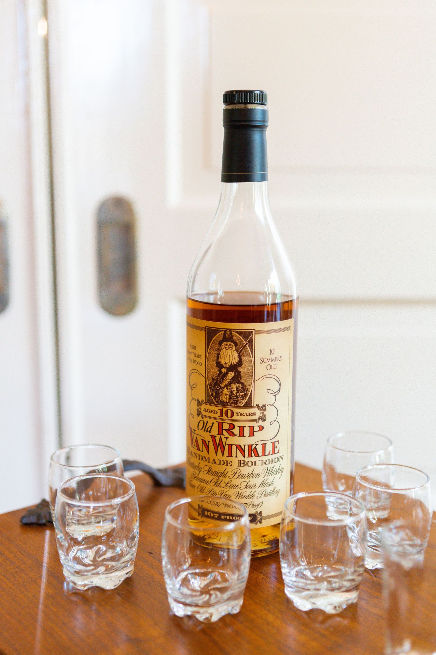 Crystal glasses and a bottle of aged whiskey sit in a row on a wooden table in soft natural lighting.