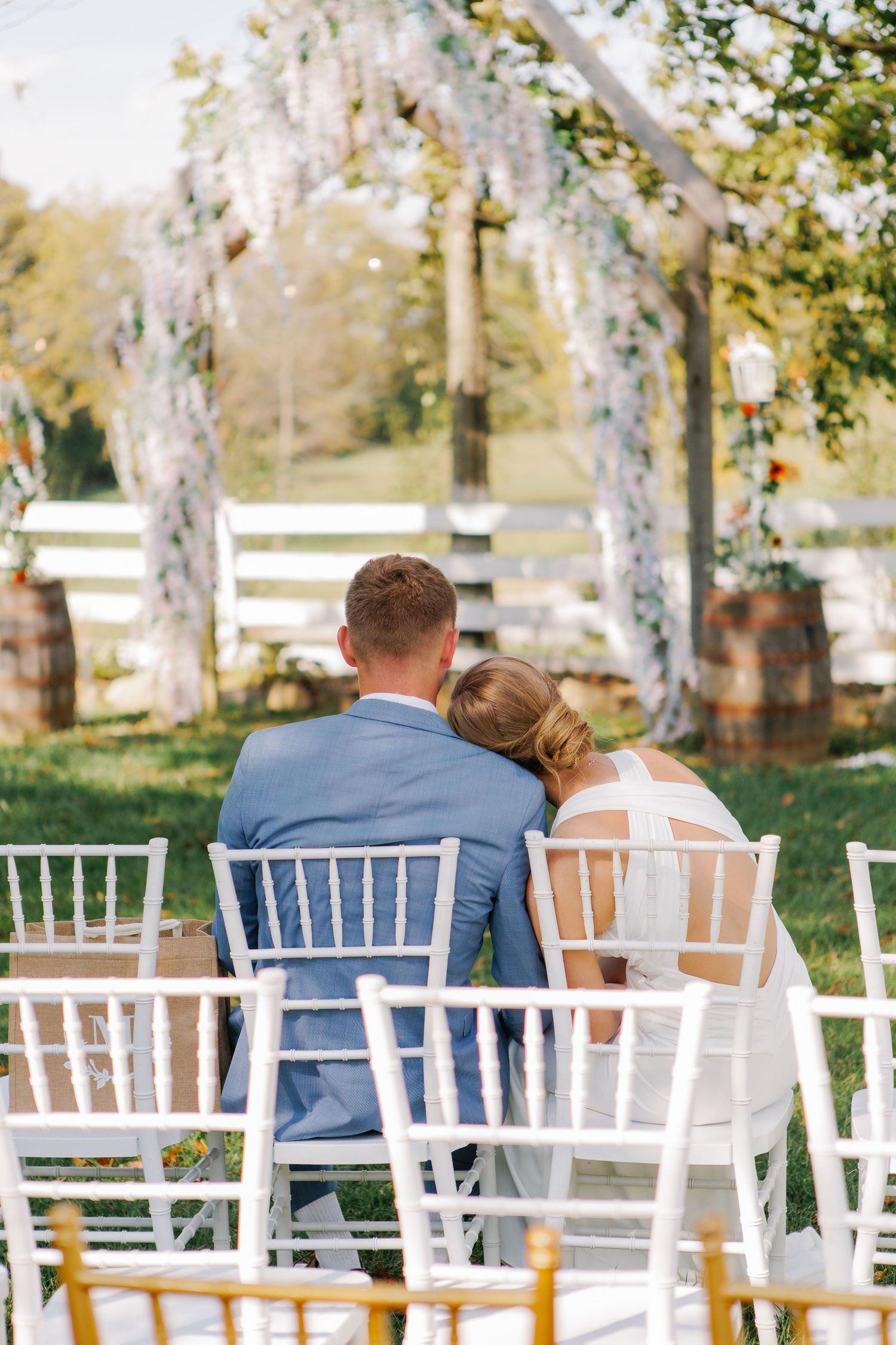Couple sitting together on white chairs under wisteria-draped wedding ceremony arch.