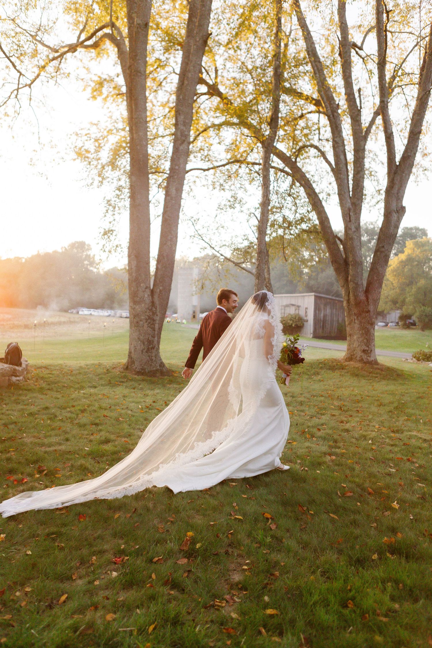 A flowing white wedding dress and veil create a romantic scene in an autumn field at sunset.