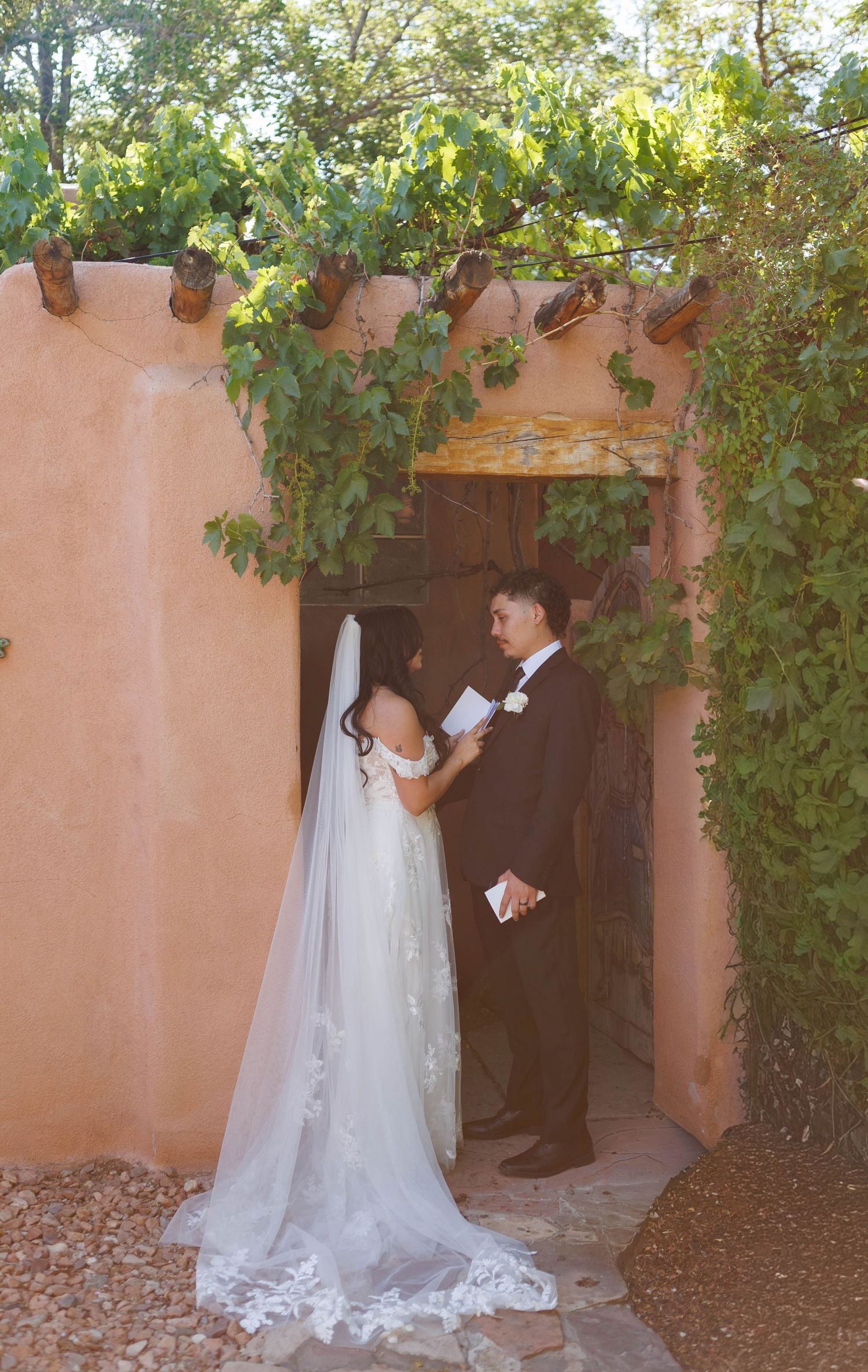Bride and groom exchange vows under a vine-covered adobe archway during an outdoor wedding ceremony.