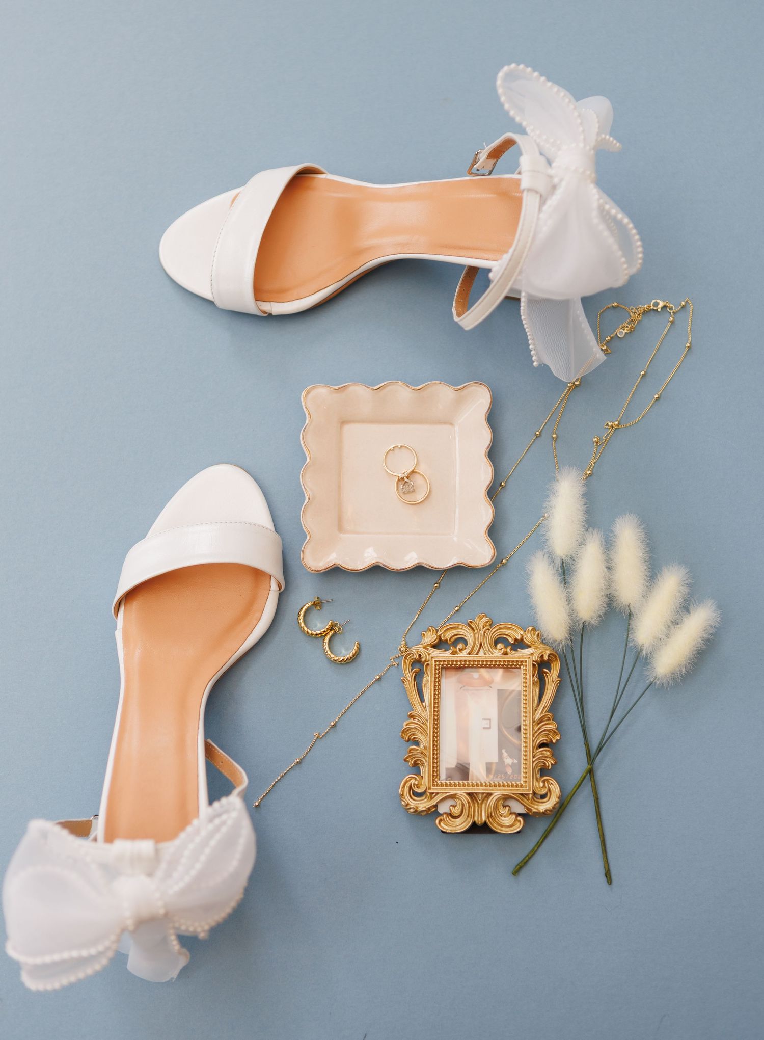 White bridal heels with bow detail beside gold rings, jewelry, dried flowers, and a vintage frame on a blue background.