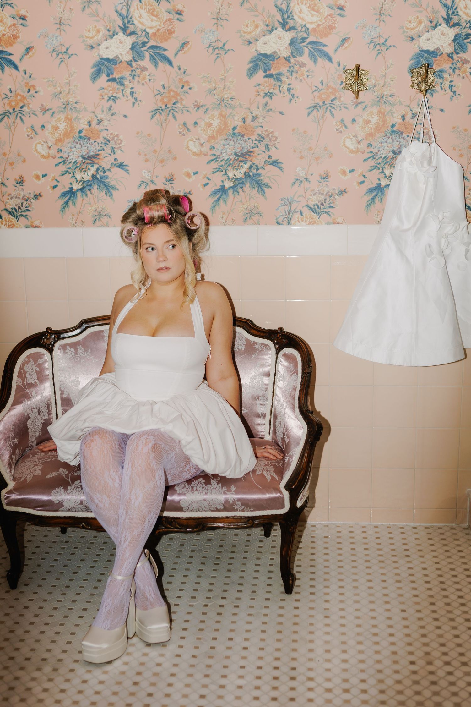 Bride in white dress and hair curlers sits on vintage pink velvet settee near floral wallpaper with wedding gown hanging nearby.