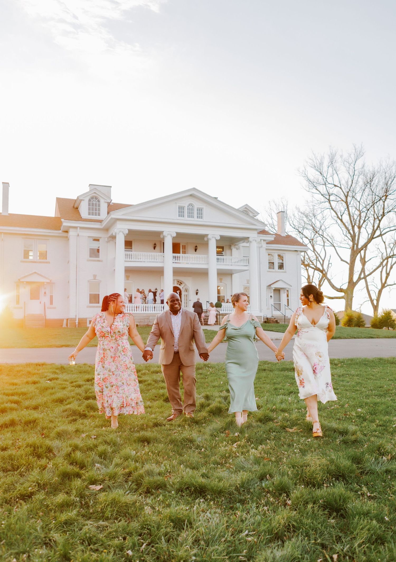 Four friends hold hands walking on green lawn before a grand white colonial mansion at golden hour sunset.