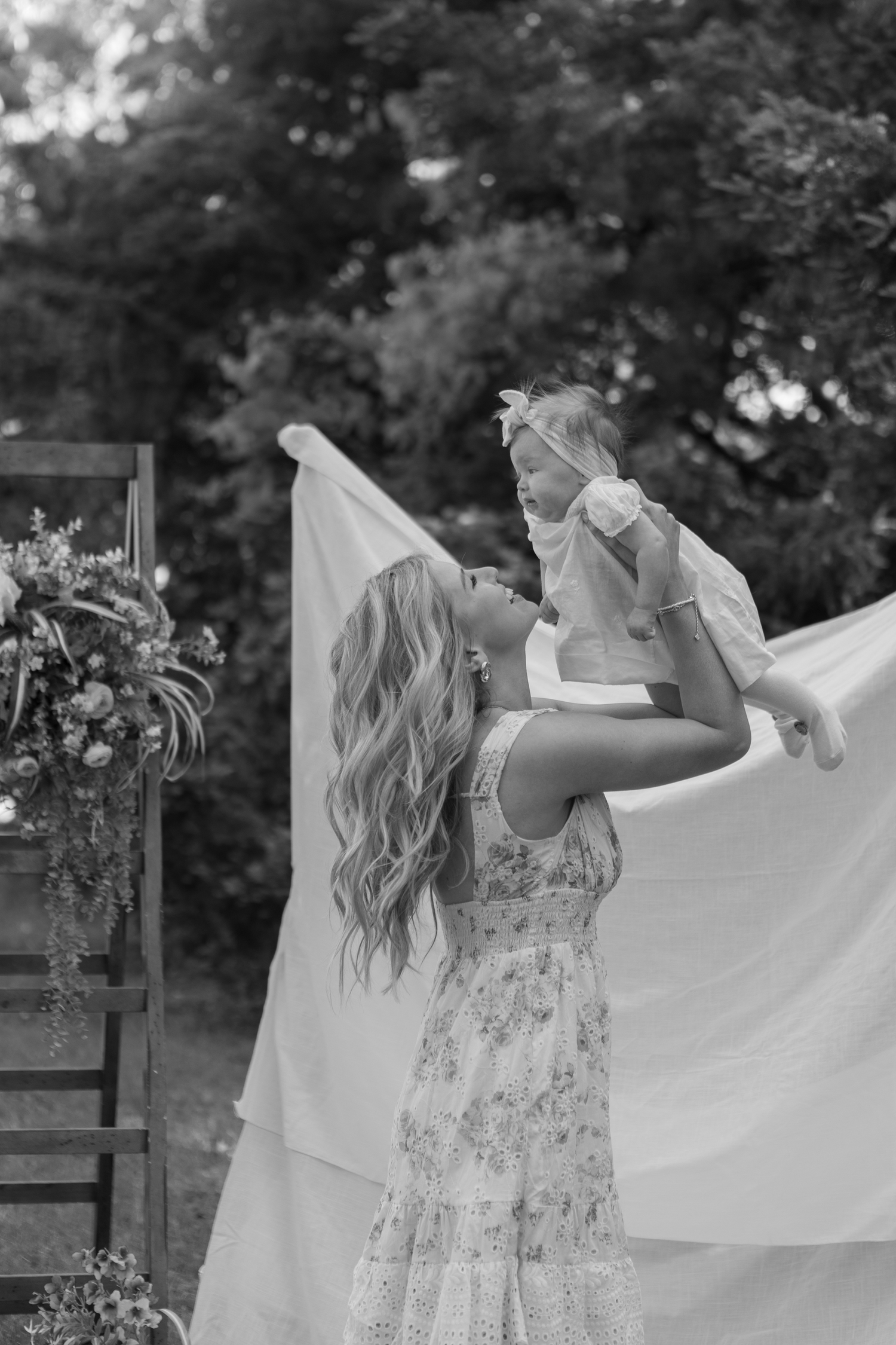 Black and white photo of a mother lifting her baby girl outdoors, with floral decor and trees in the background.