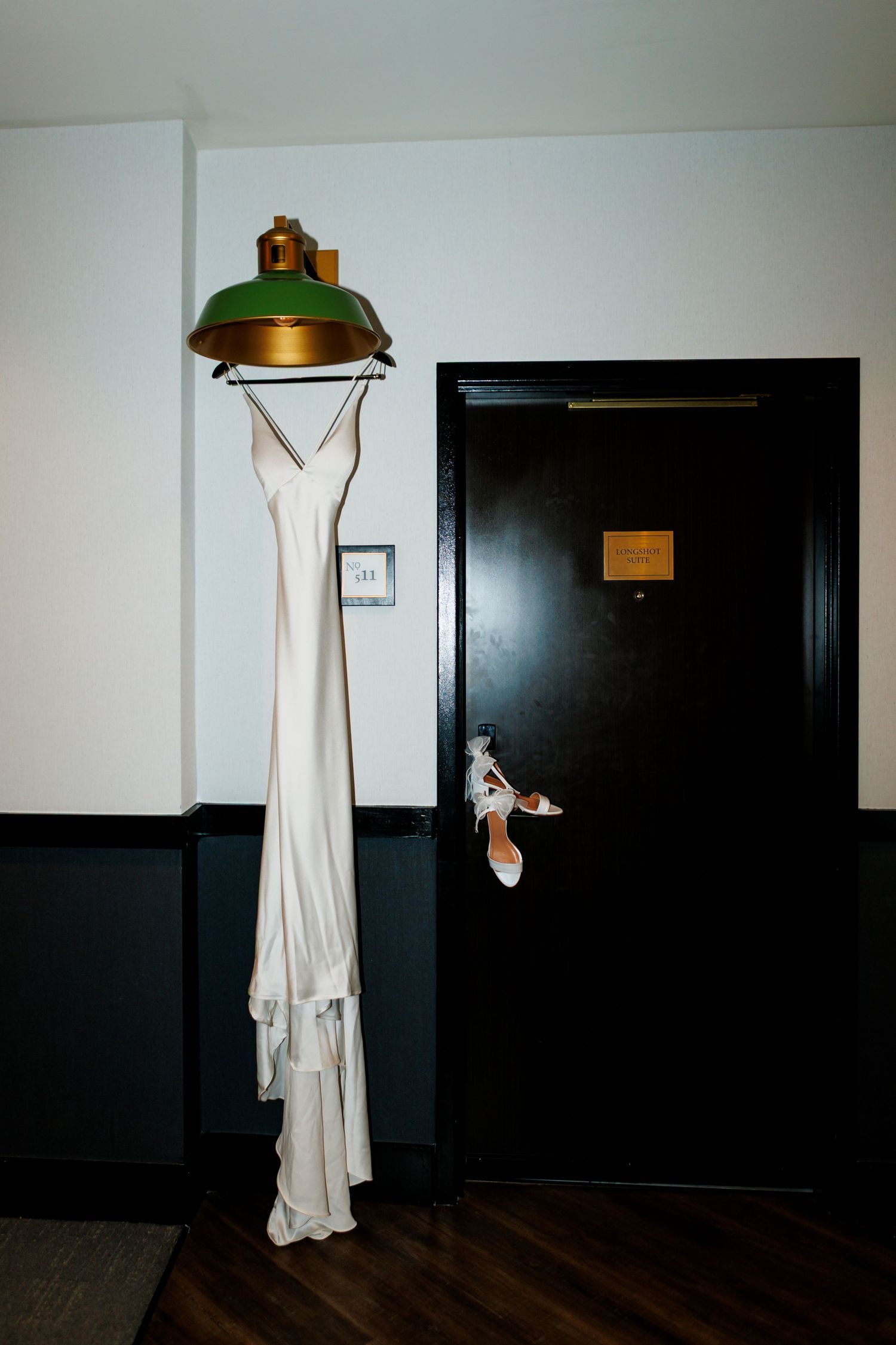 White wedding dress hanging on a vintage green lamp fixture beside a dark door with bridal heels.