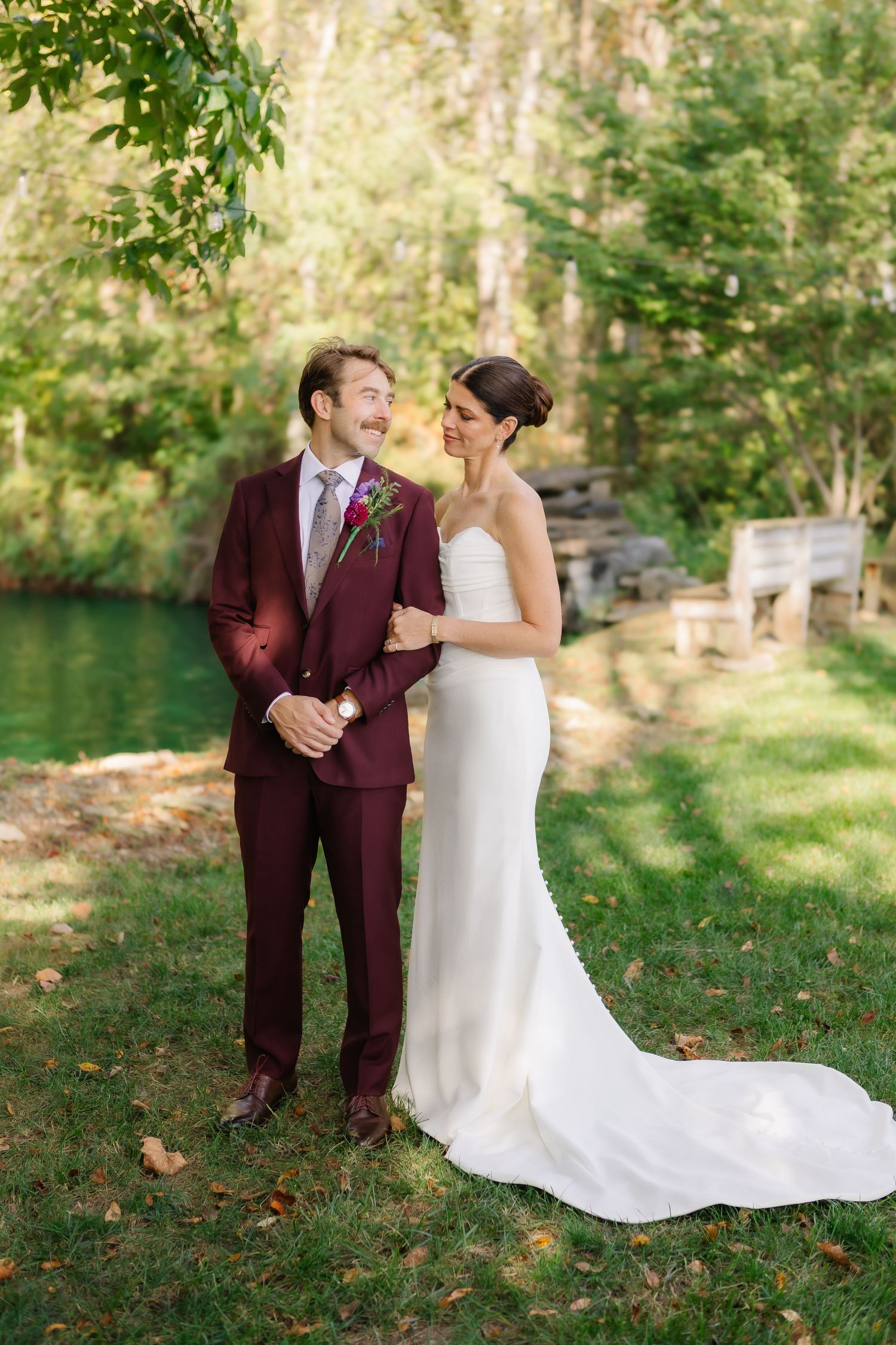 A romantic outdoor wedding scene with a couple in burgundy suit and white dress embracing on a grassy lawn.