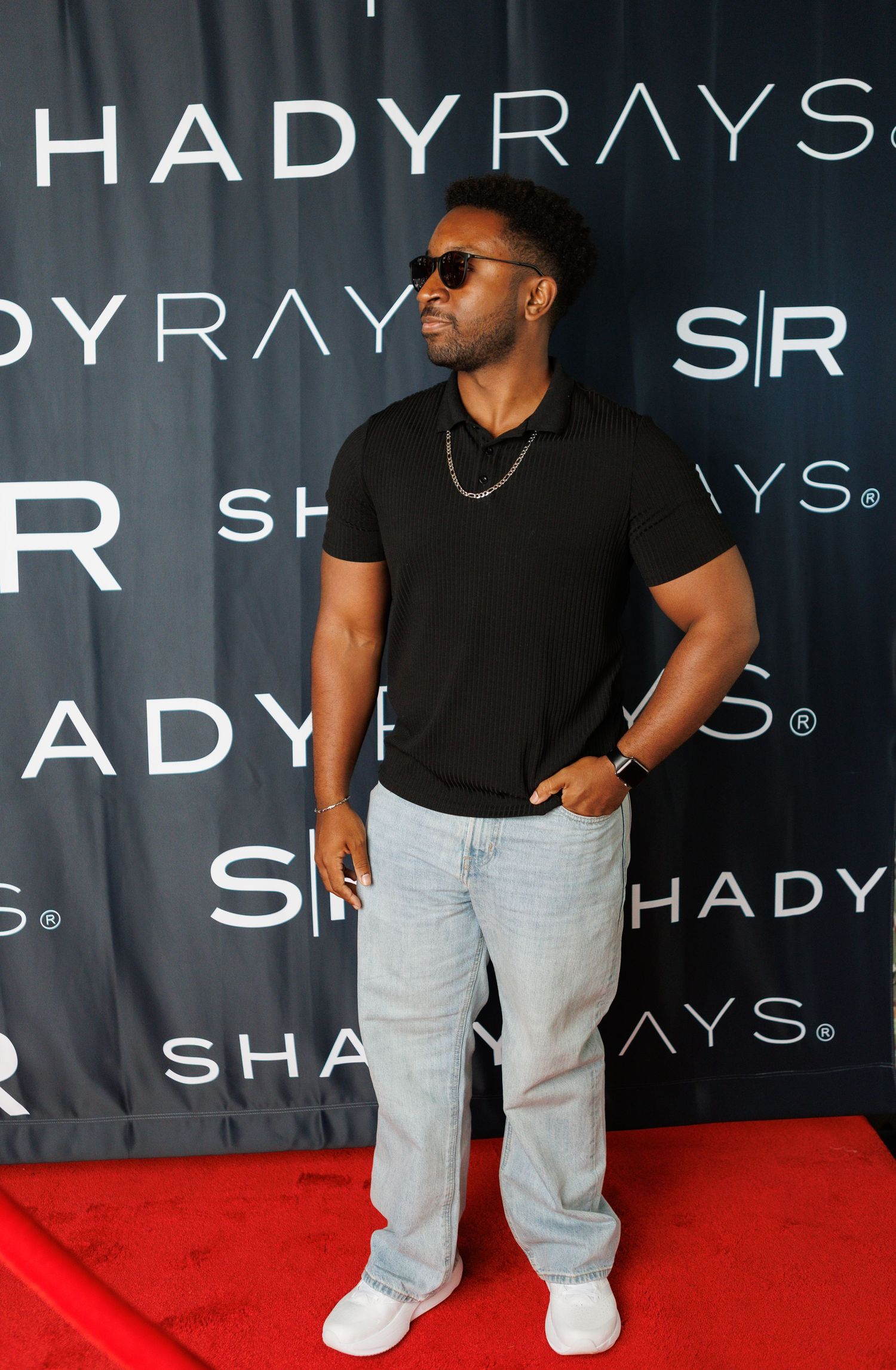 Man in black polo shirt and jeans posing on red carpet at Shady Rays sunglasses event.