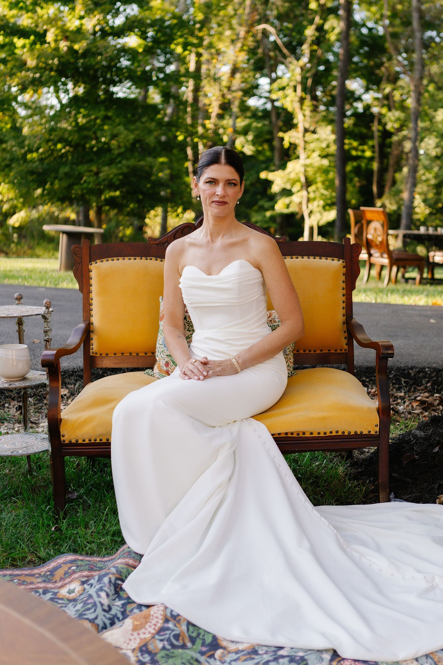 A bride in a classic white wedding gown poses elegantly on a vintage yellow couch outdoors in a garden setting.