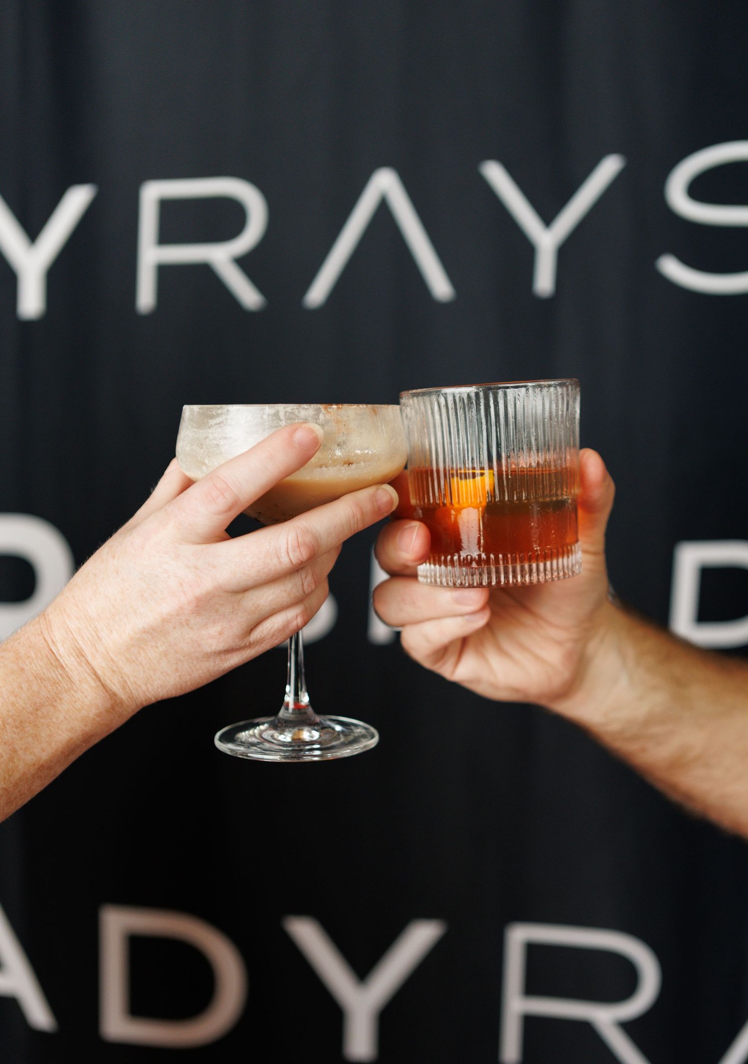 Two hands clinking cocktail glasses in front of a dark ADYRAYS branded backdrop.