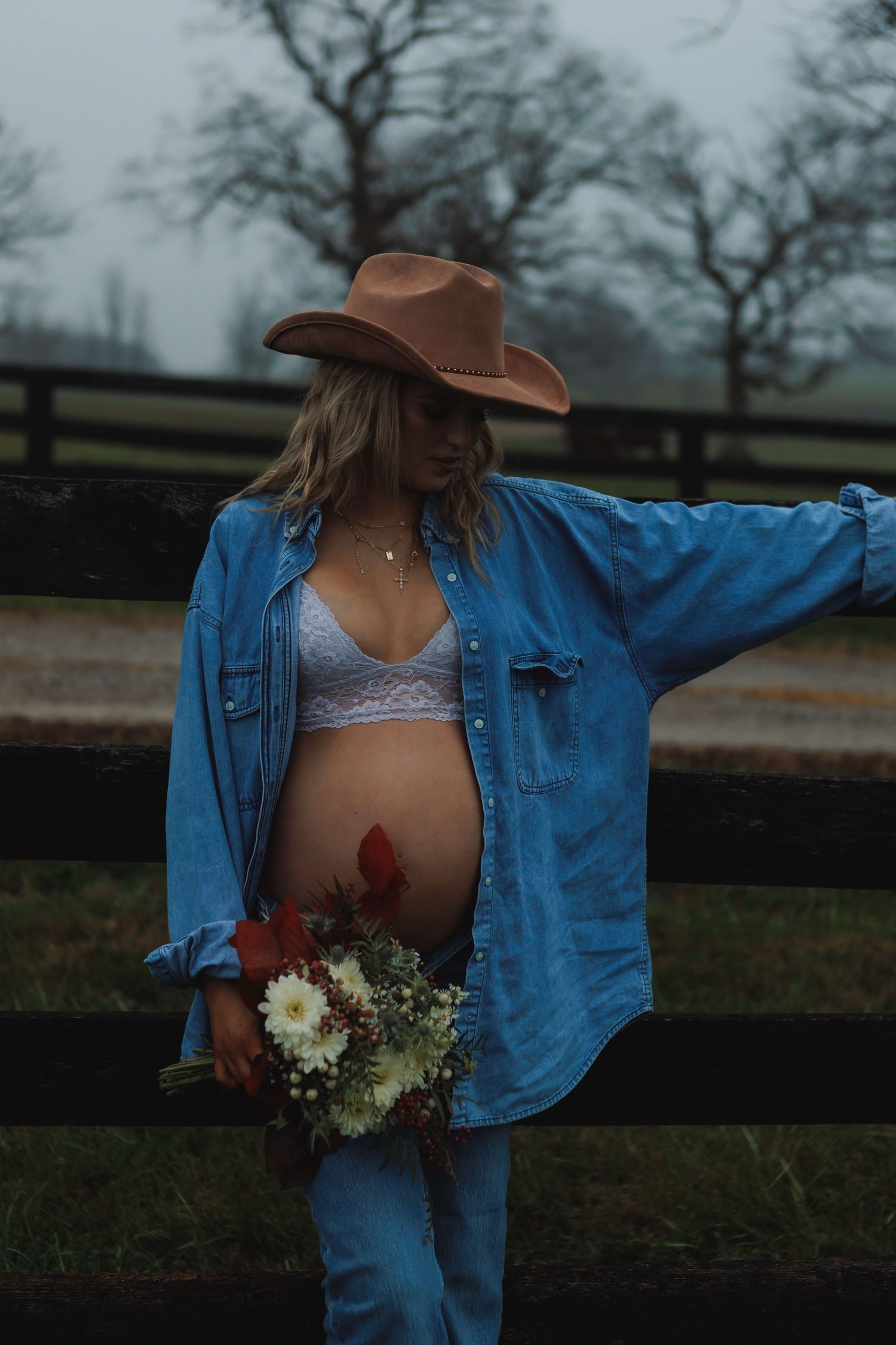 Person in denim and cowboy hat poses against wooden fence with wildflowers.