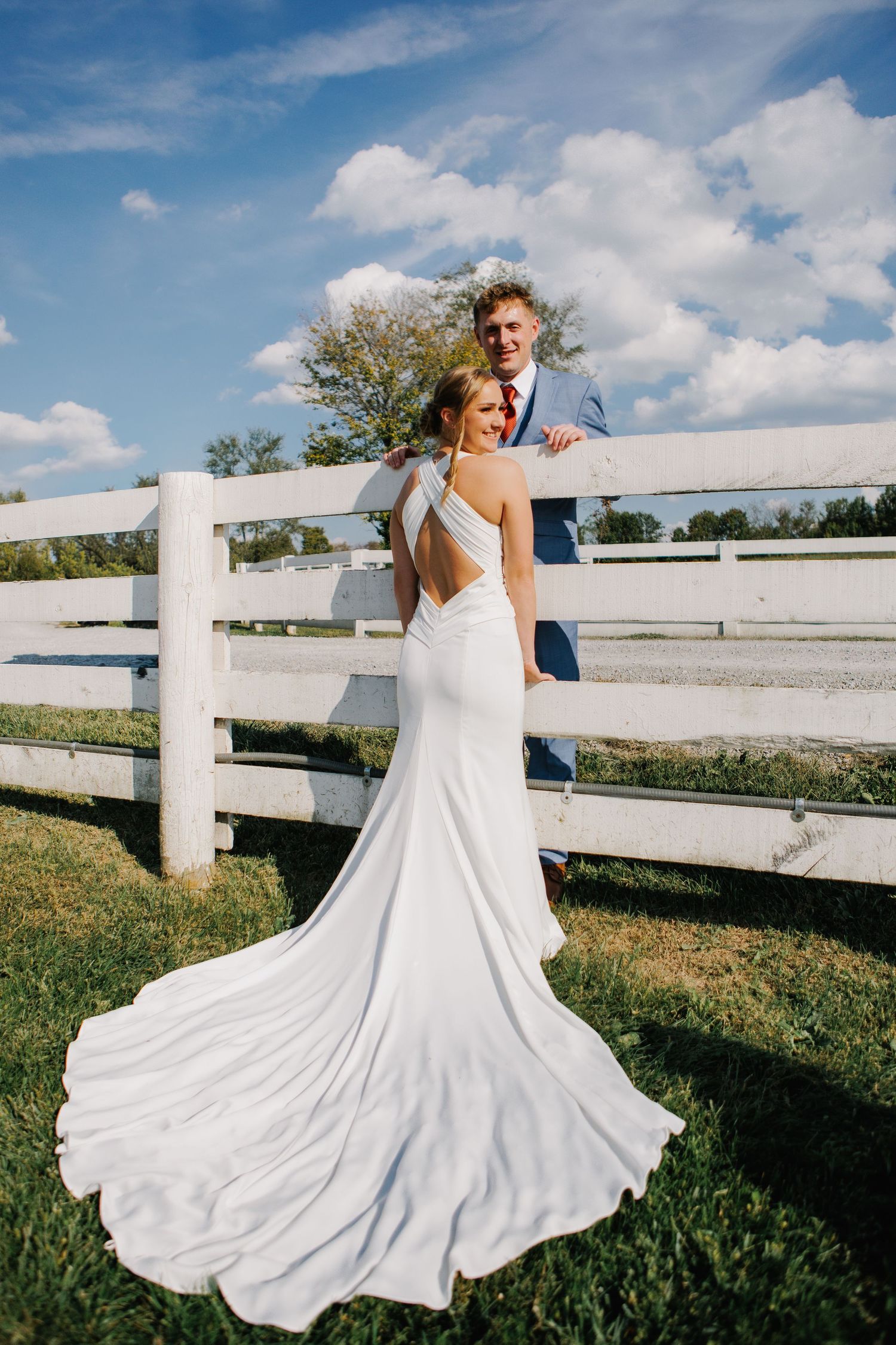 Bride in flowing white dress twirling against rustic white fence on sunny day.