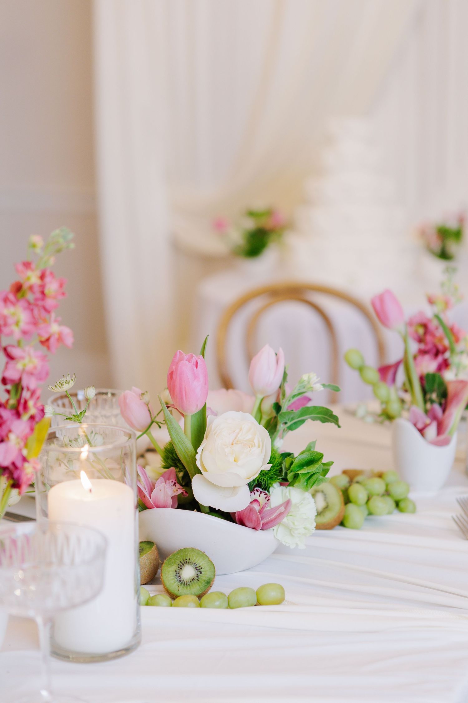Wedding reception table decorated with pink tulips, white roses, candles, grapes, and kiwi fruit centerpieces.