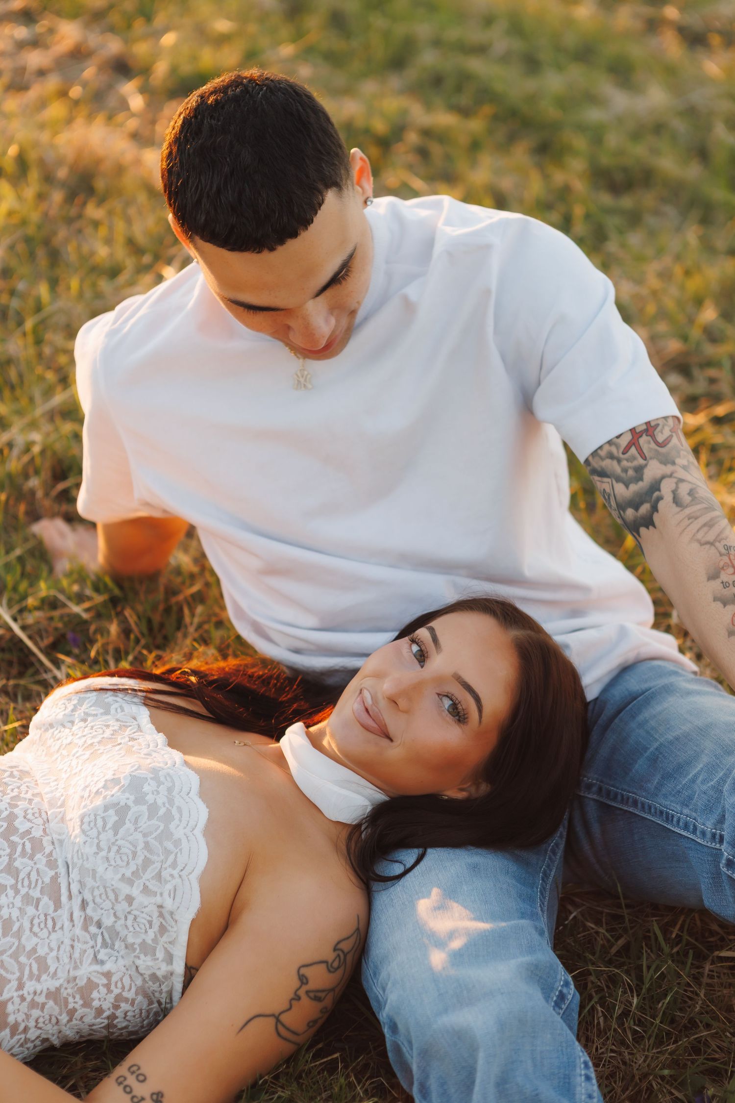 A tattooed couple relaxes on grass at golden hour, woman lying in mans lap smiling at camera in lace top and jeans.