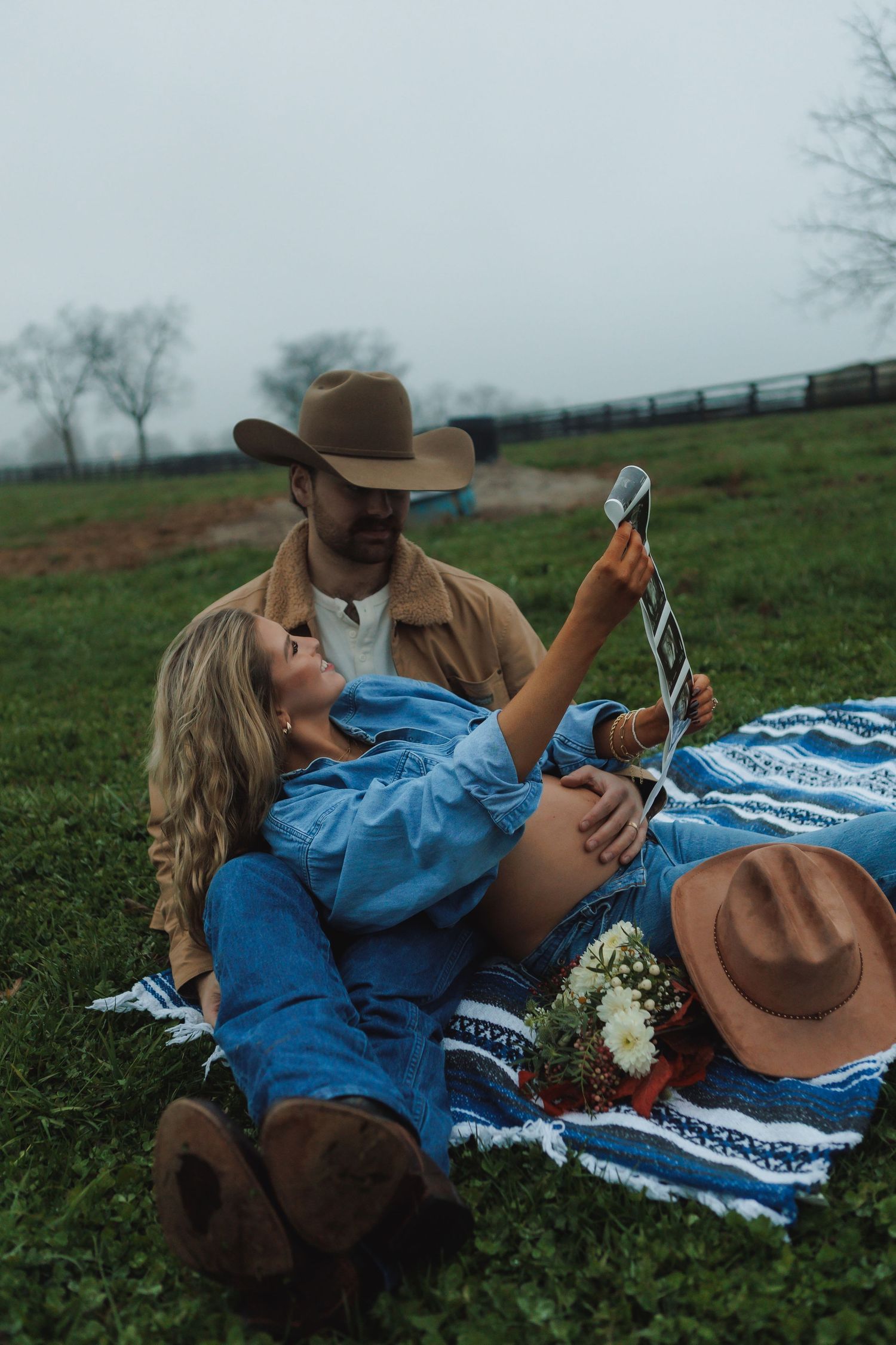 A couple in western wear relaxes on a picnic blanket in a grassy field.