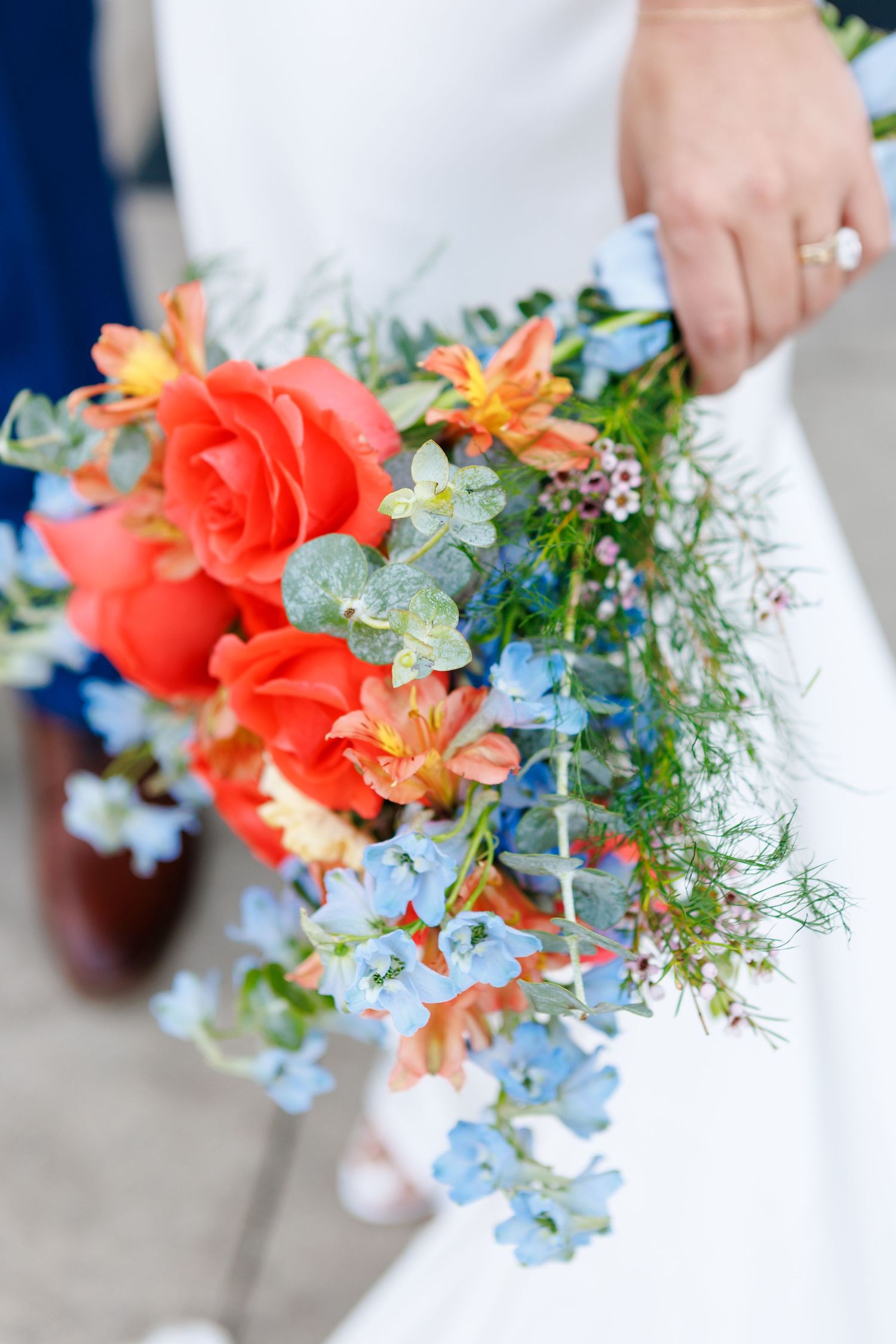 Bride holding a vibrant wedding bouquet with red roses, orange lilies, and blue wildflowers on her special day.