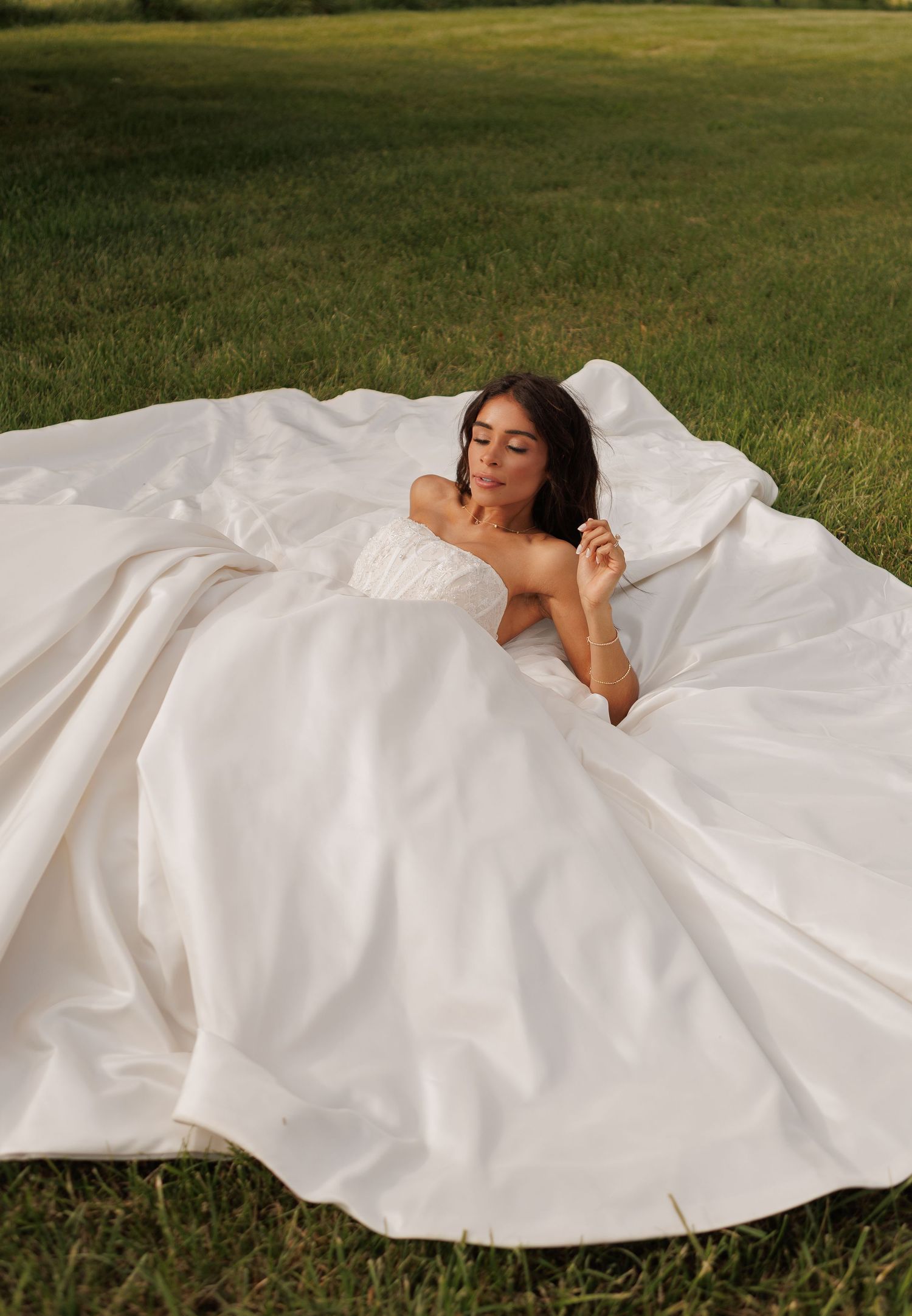 Bride in a flowing white strapless ball gown laying on green grass, with a dramatic oversized skirt spread around her.